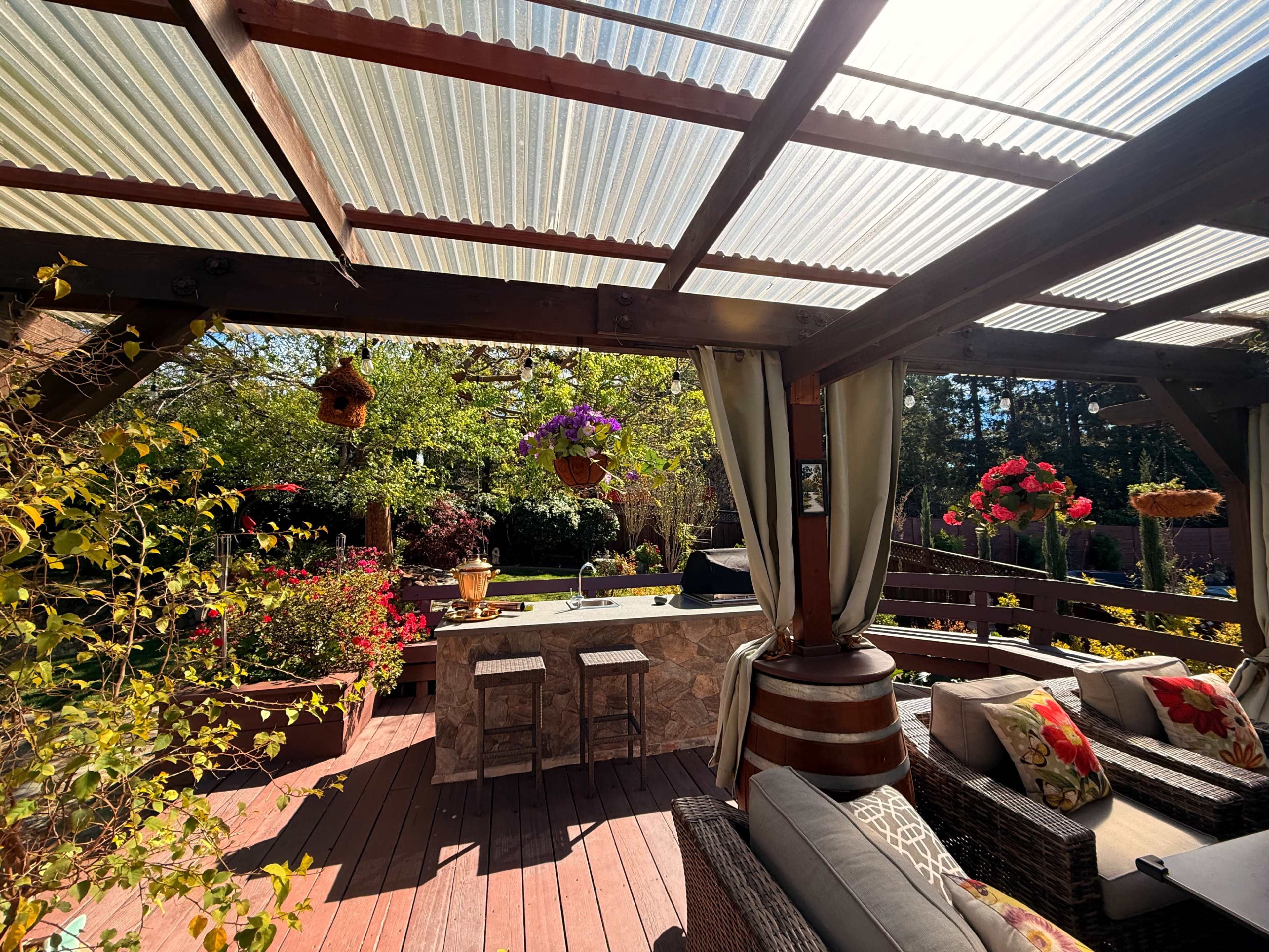 The image shows a backyard patio with a bar area, surrounded by colorful flowers and plant pots, and a wooden deck with comfortable seating.