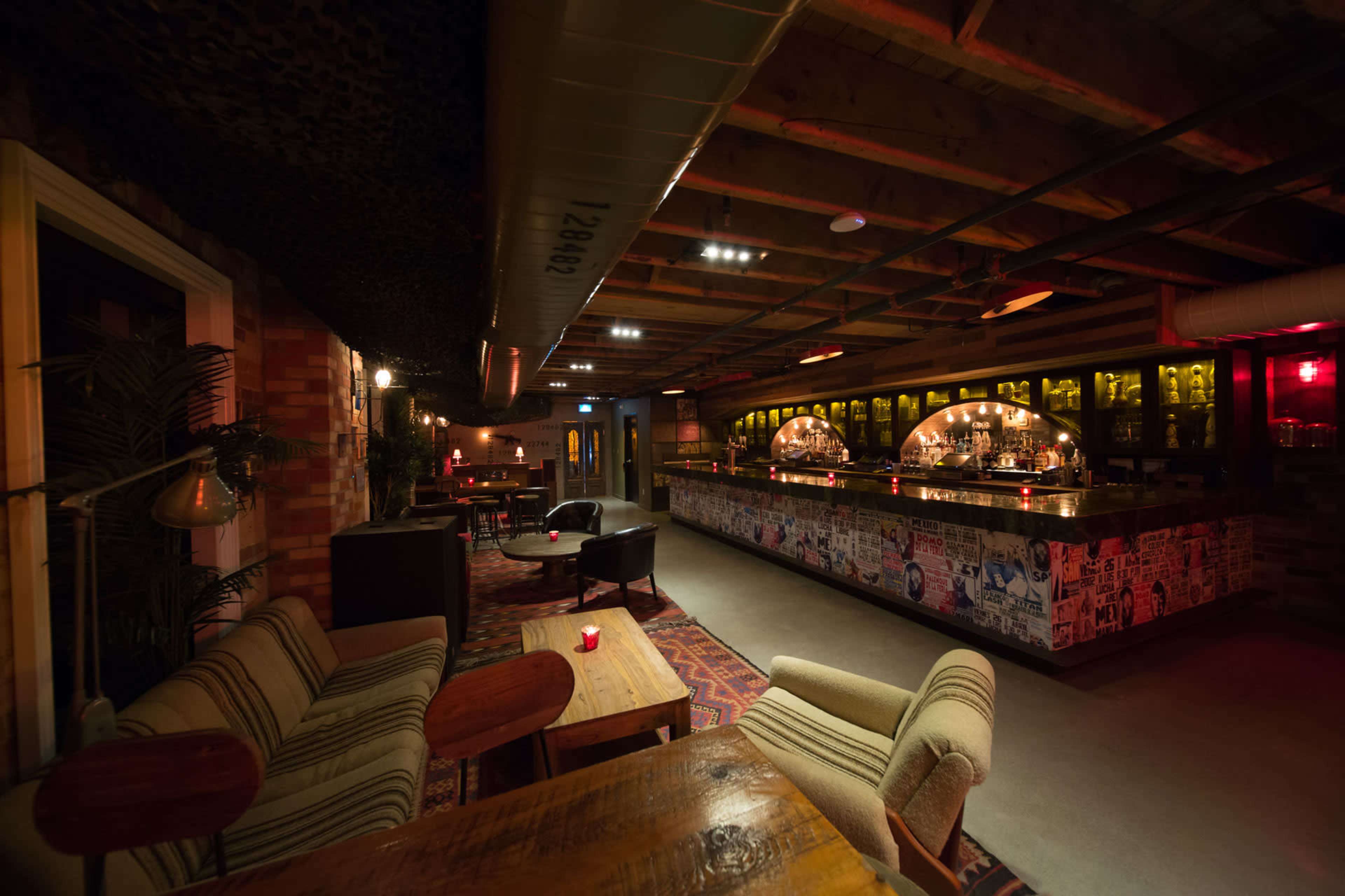 The image shows a dimly lit bar with a long counter, wooden tables, and upholstered chairs, featuring a stylish decor and a patterned carpet.