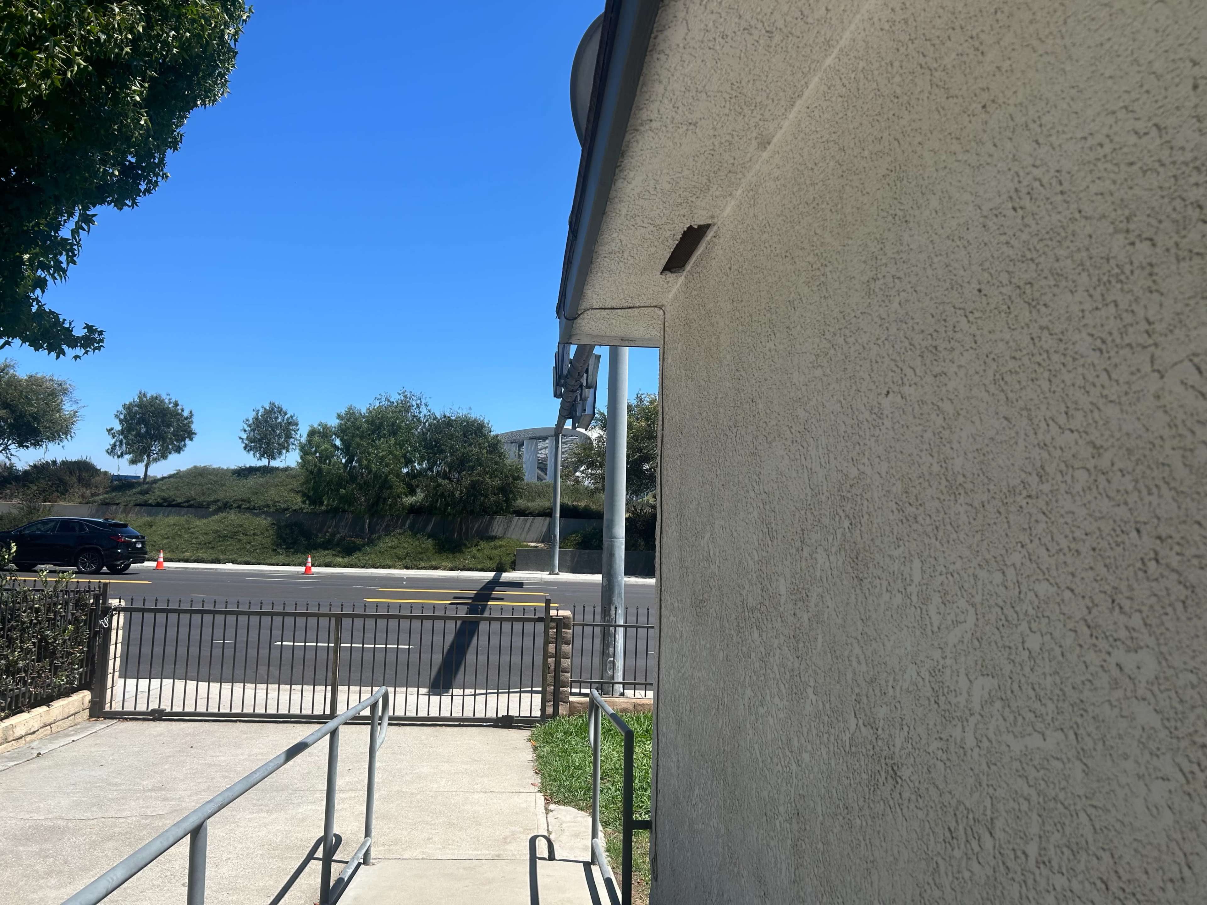 The image shows a view of a sidewalk leading to a building, with a parking lot and street visible in the background under a clear blue sky.