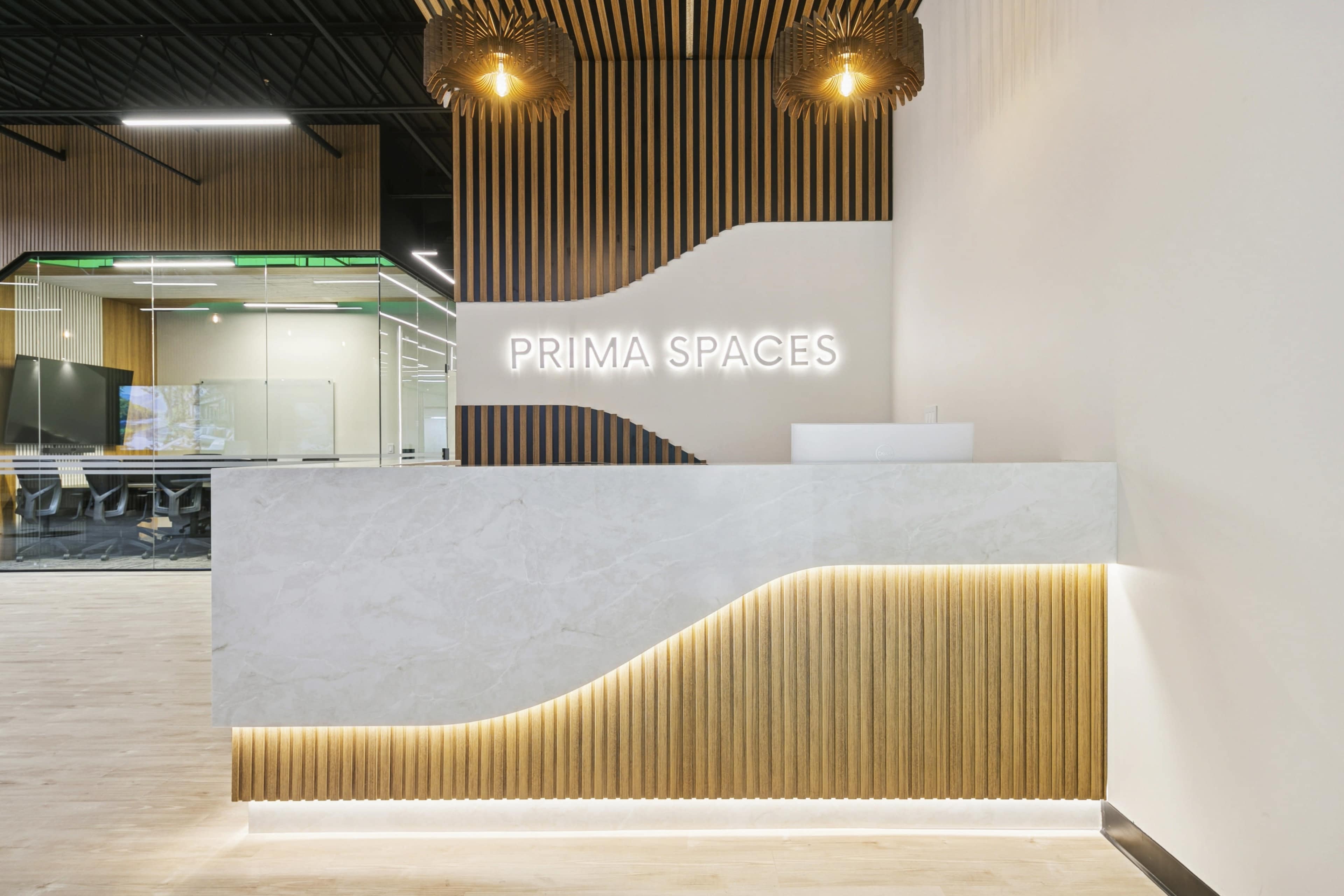 The image shows a modern reception area with a sleek white marble front desk, wooden paneling, and illuminated signage that reads "PRIMA SPACES."