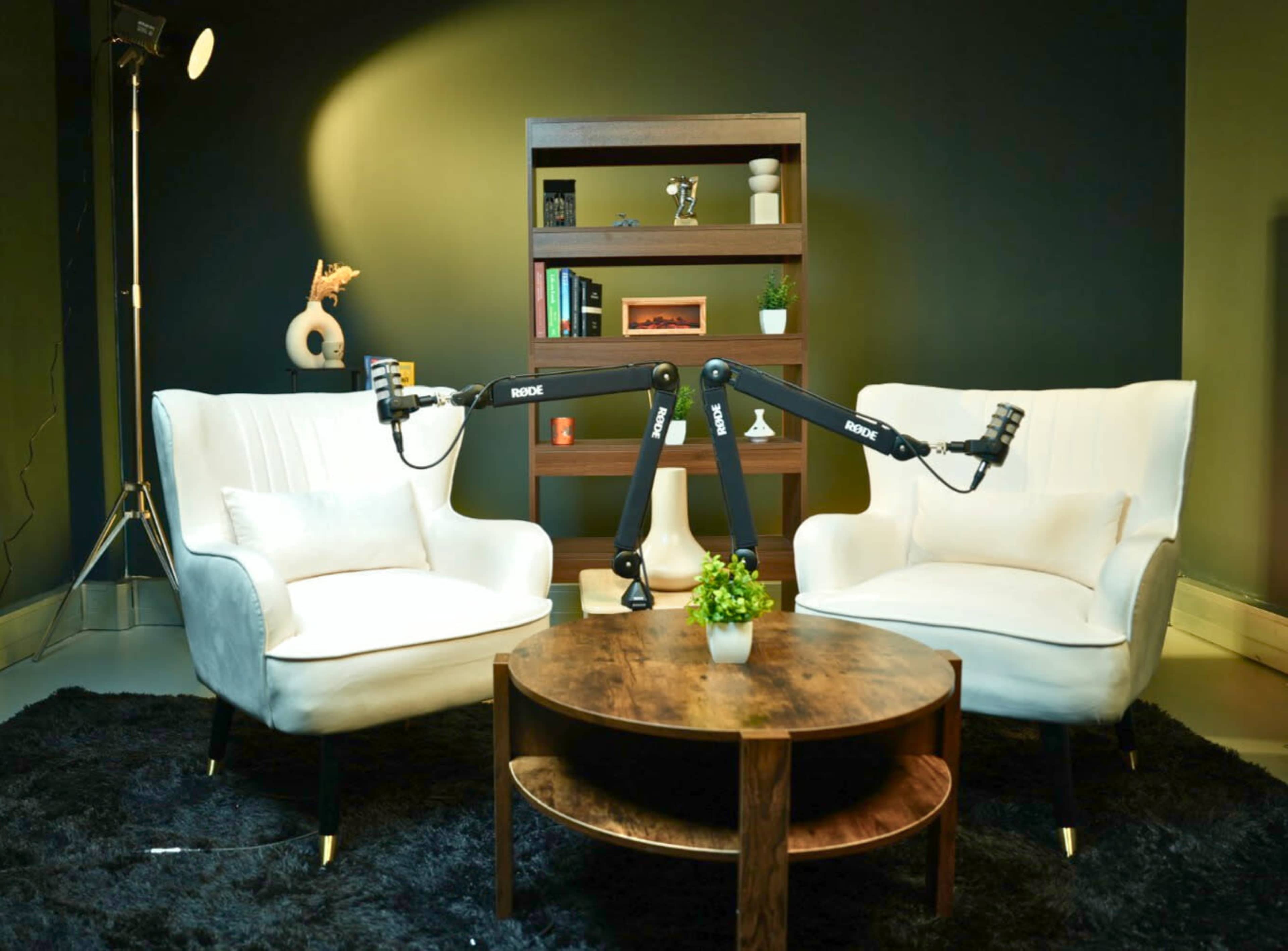 The image features two white chairs facing each other over a round wooden coffee table, with a dark green wall and a bookshelf in the background.