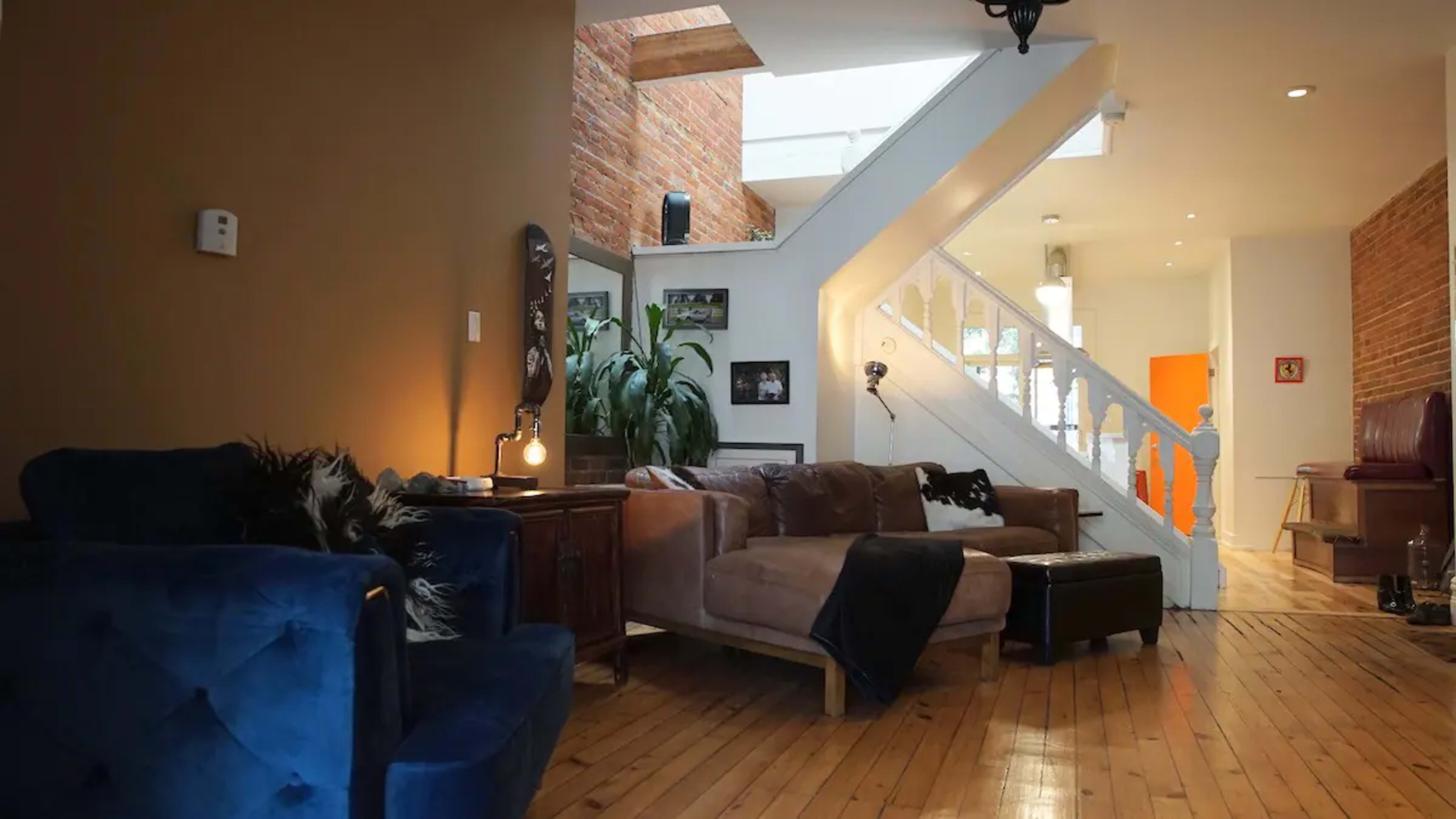 The image shows a modern living room with wooden flooring, a staircase leading to a brighter level, and a mix of plush furniture and decorative elements.