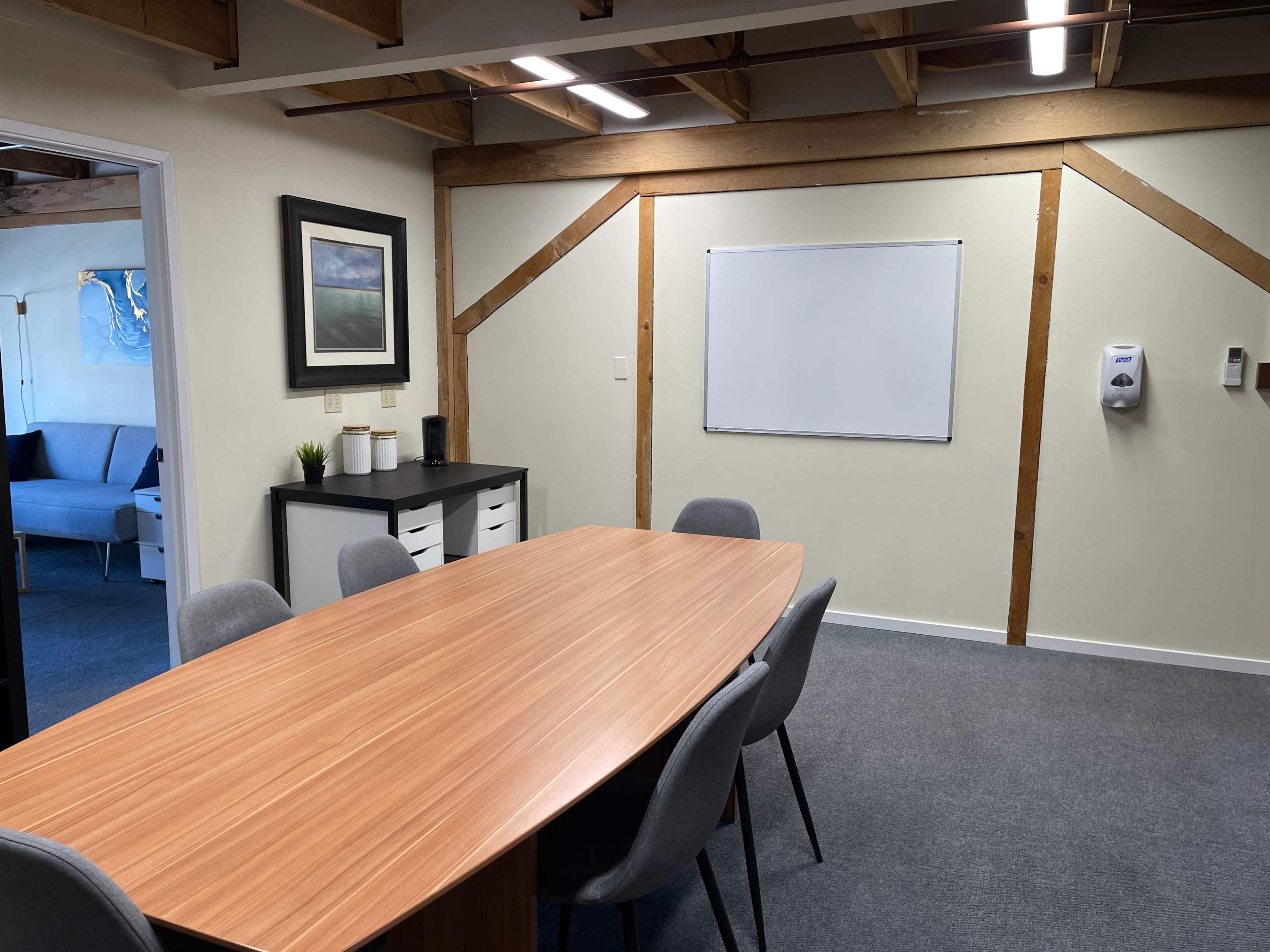 A conference room with a wooden oval table surrounded by gray chairs, a whiteboard on the wall, and a cabinet with drawers.