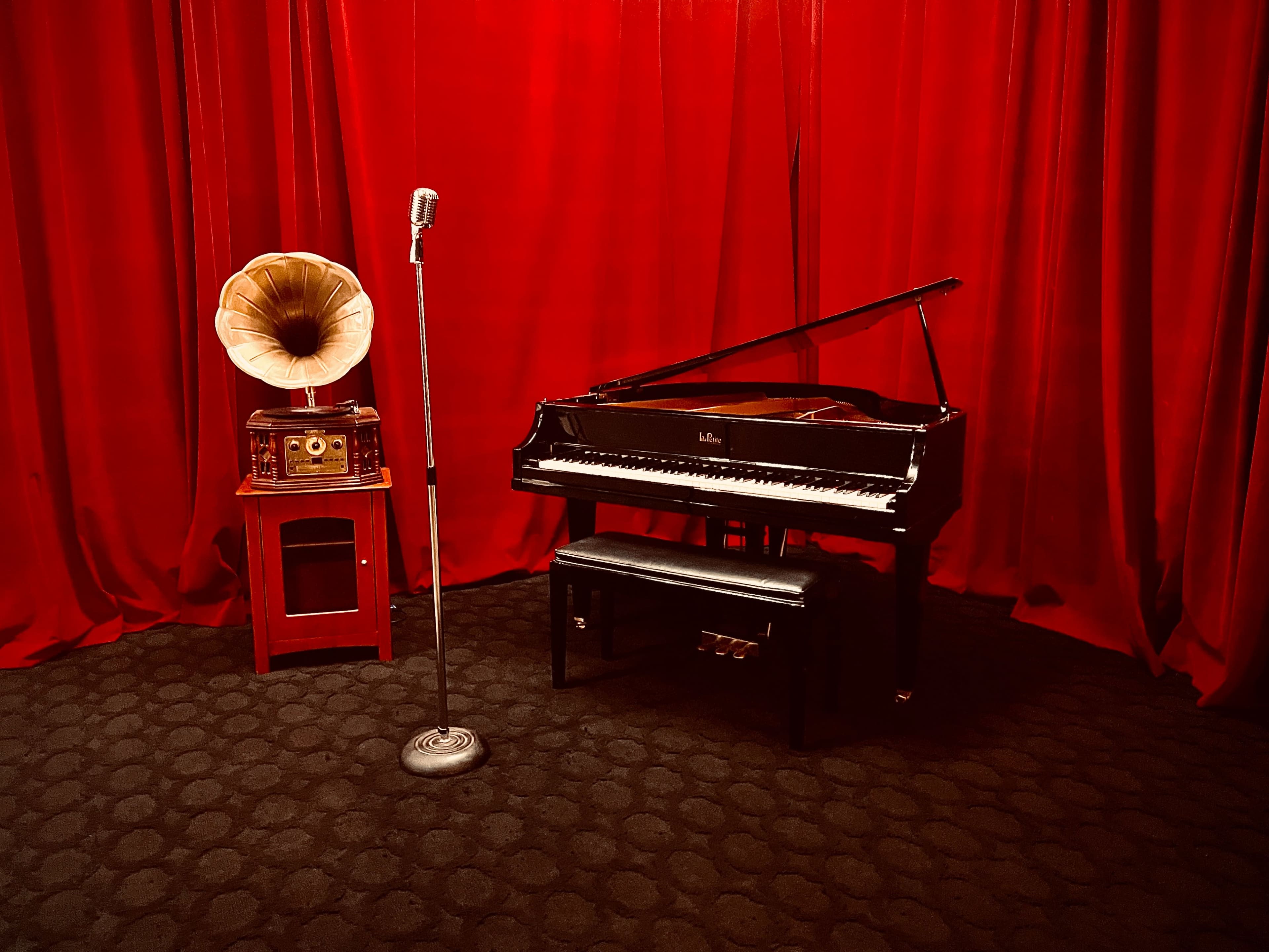 A grand piano and a vintage gramophone are set against a red velvet backdrop, with a microphone on a stand nearby.