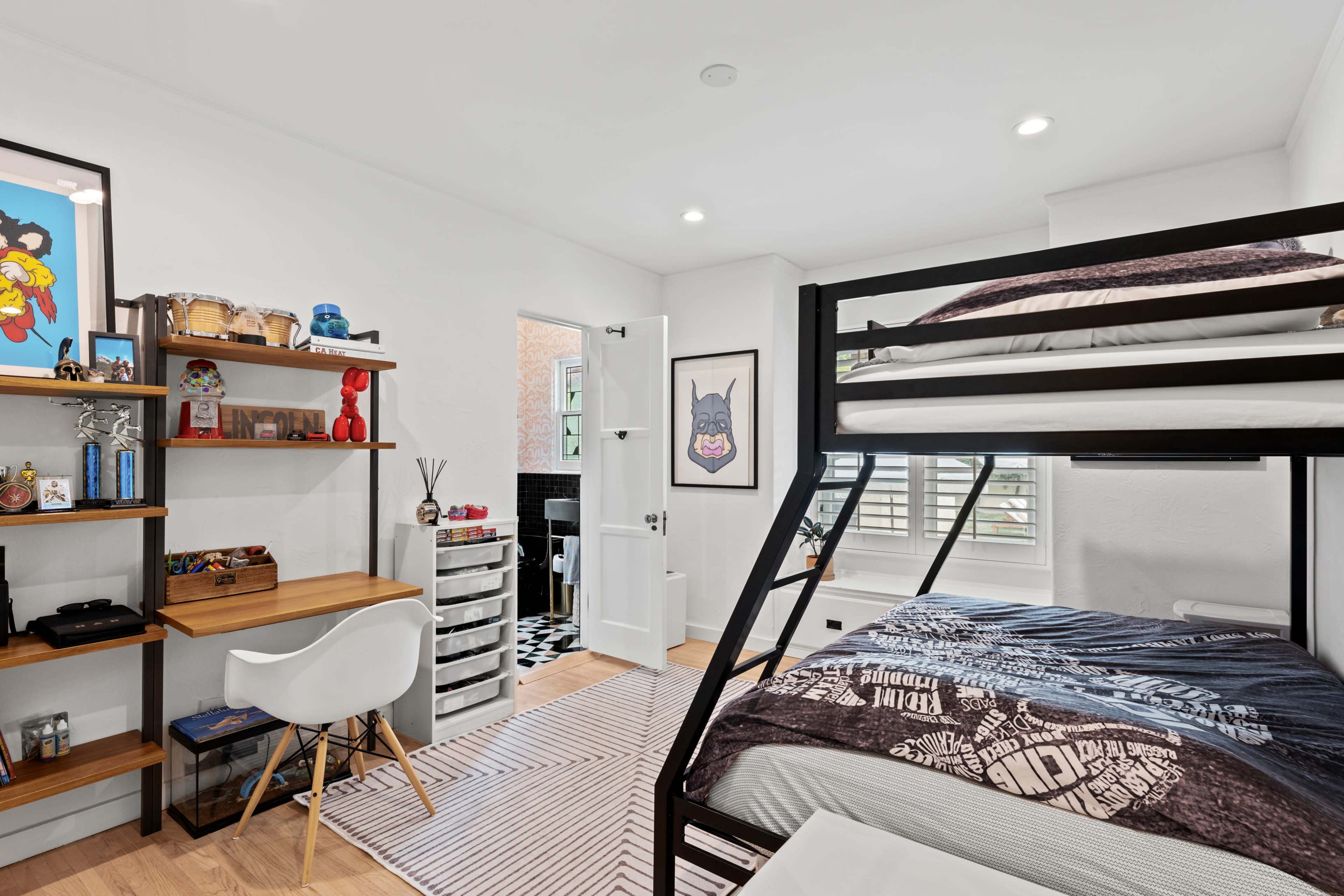 A modern bedroom features a bunk bed, a wooden desk with a chair, and a door leading to a small bathroom.
