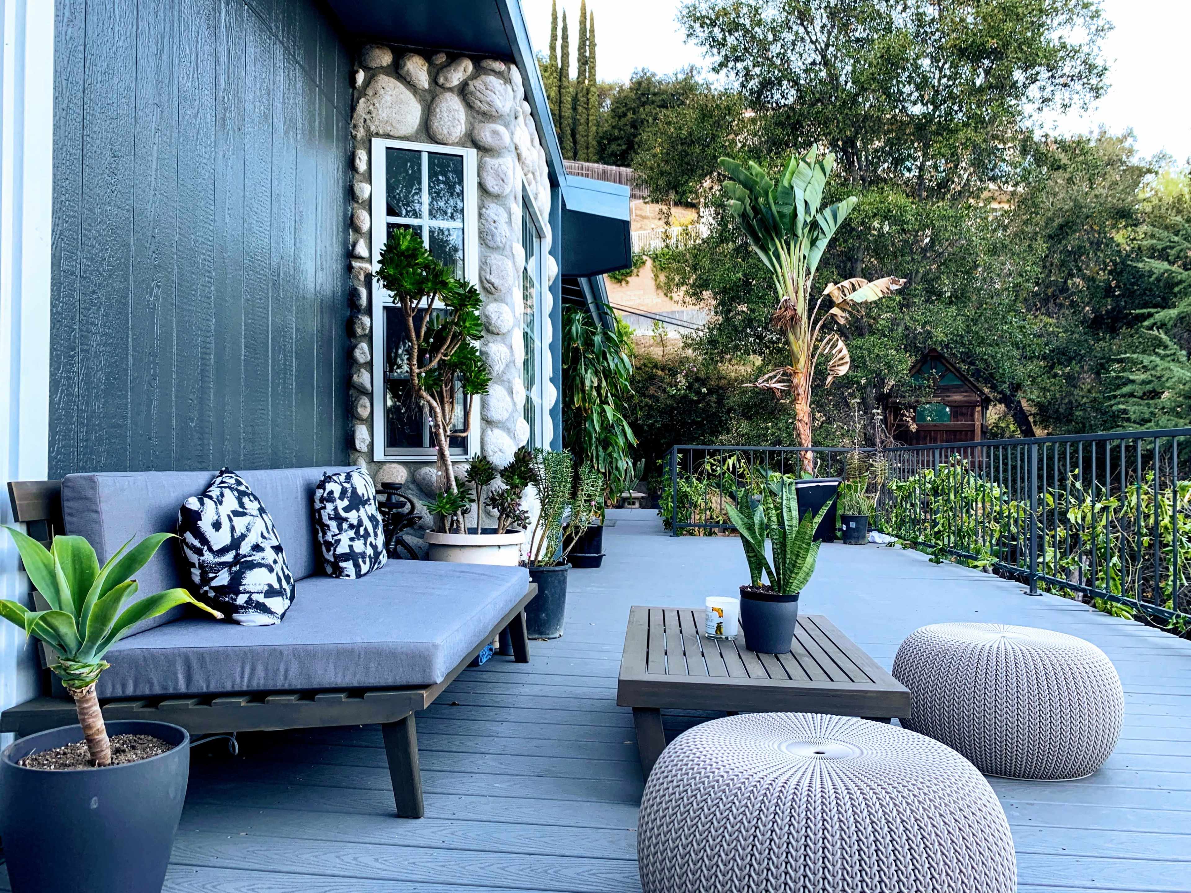 A modern patio area with a gray sofa adorned with patterned pillows, surrounded by potted plants and a low wooden coffee table, set against a backdrop of greenery.