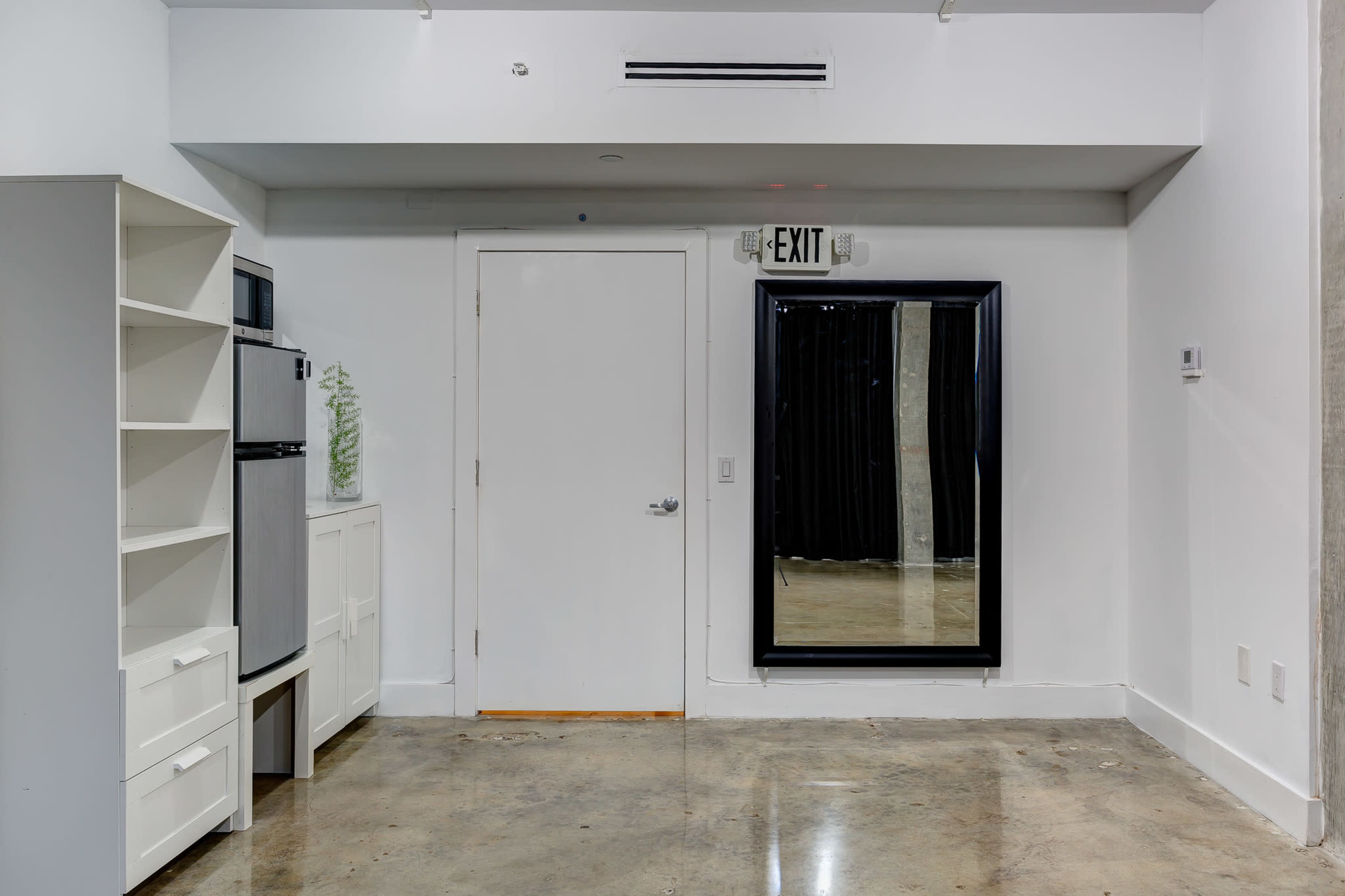 A simple room with a refrigerator, a white cabinet, a door, a large mirror, and an exit sign, all set against a polished concrete floor.