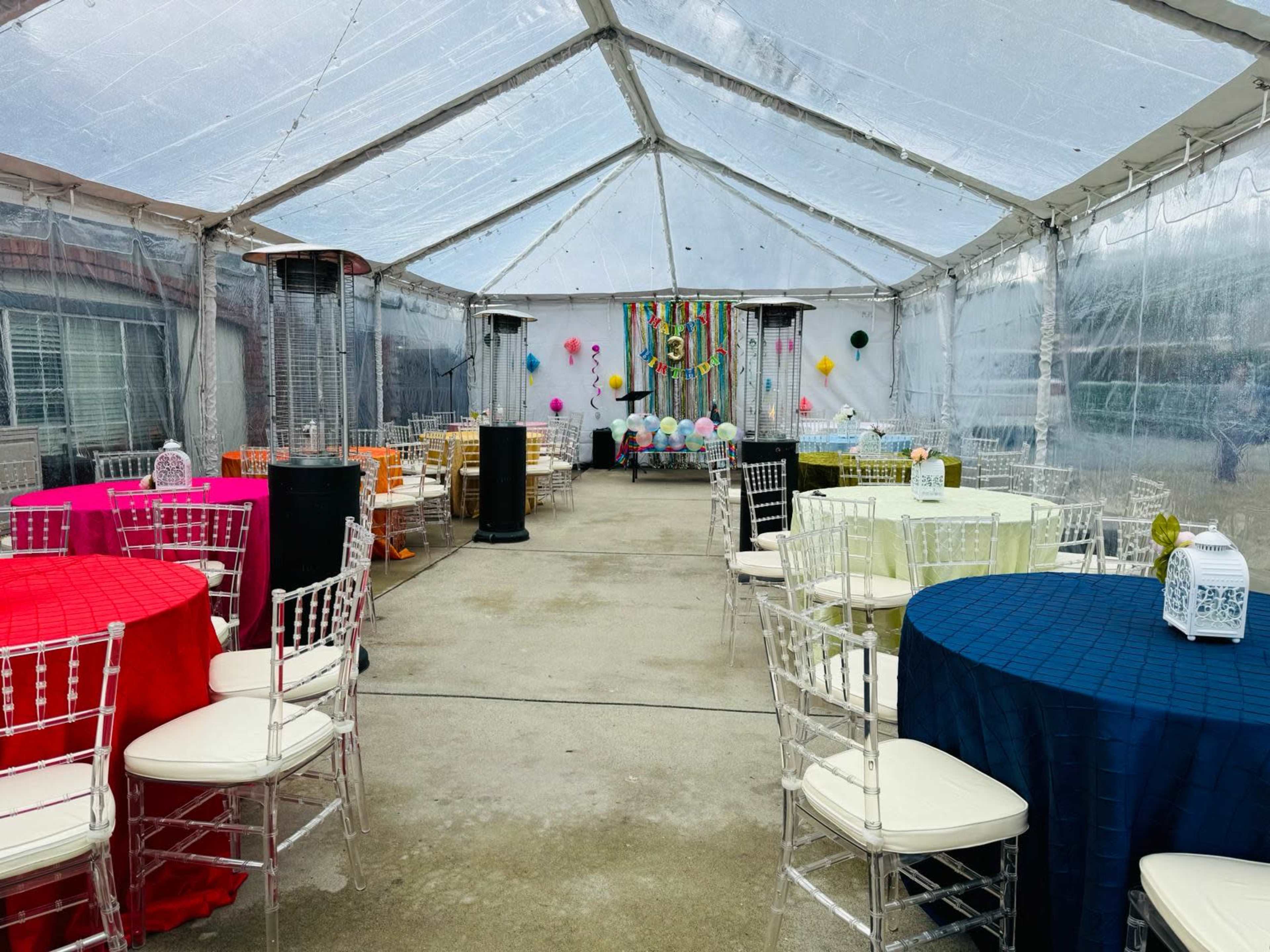 A tented event space features several tables covered in different colored cloths, with decorative elements and balloons in the background.