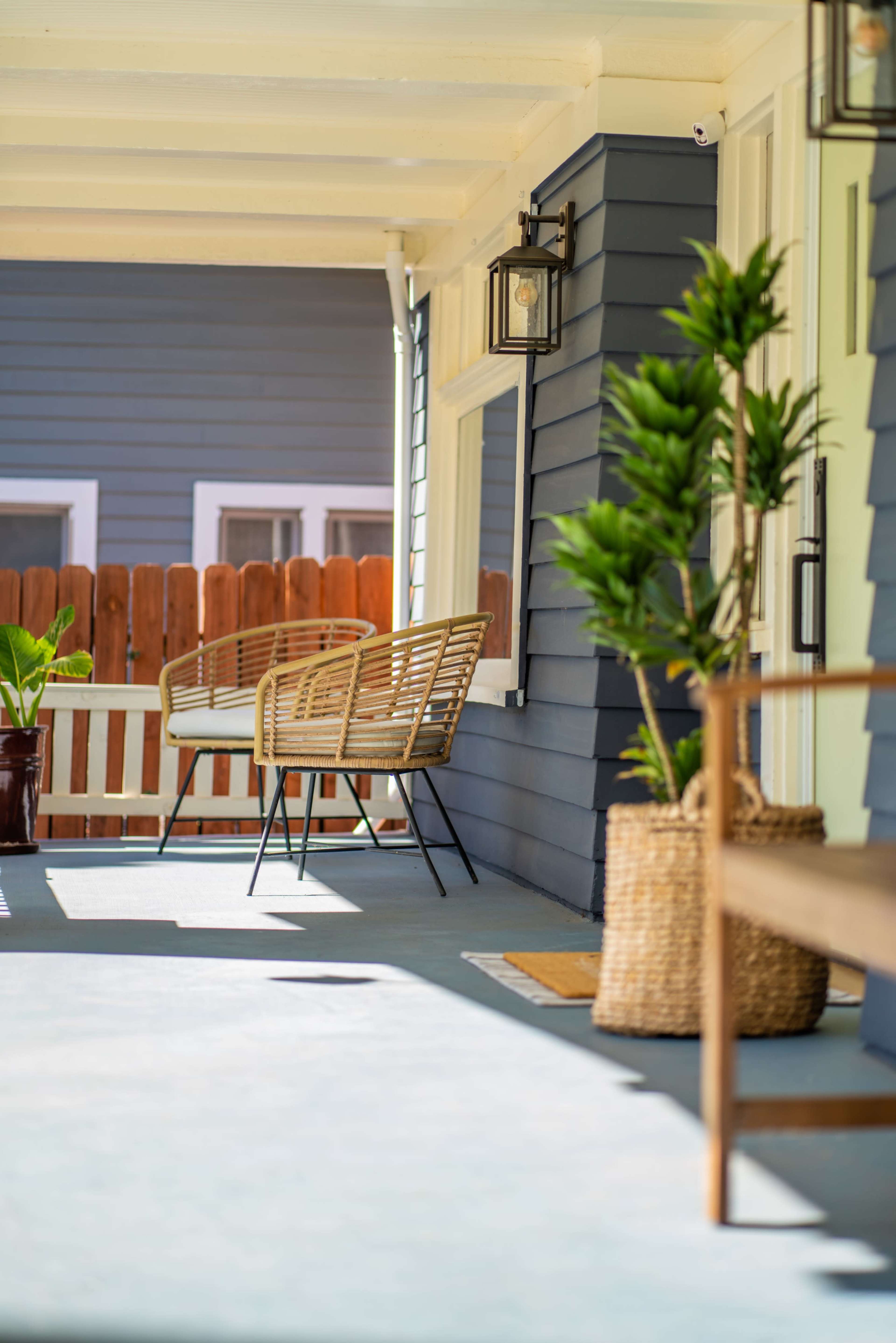 A porch with a rattan bench, potted plants, and a wooden side table against a blue and white house.