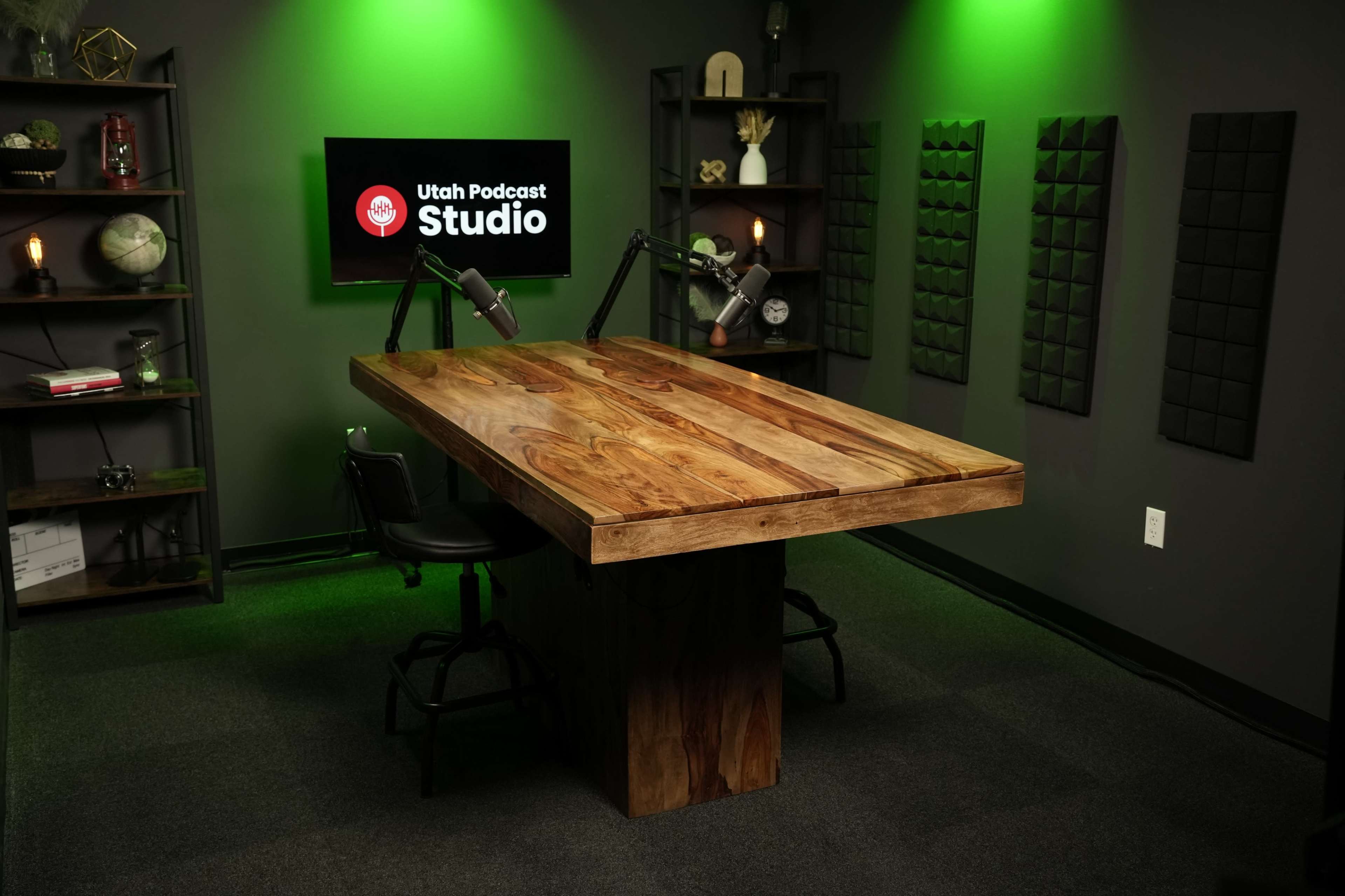 The image shows a podcast studio featuring a large wooden table, two microphone stands, and green lighting against a dark backdrop.