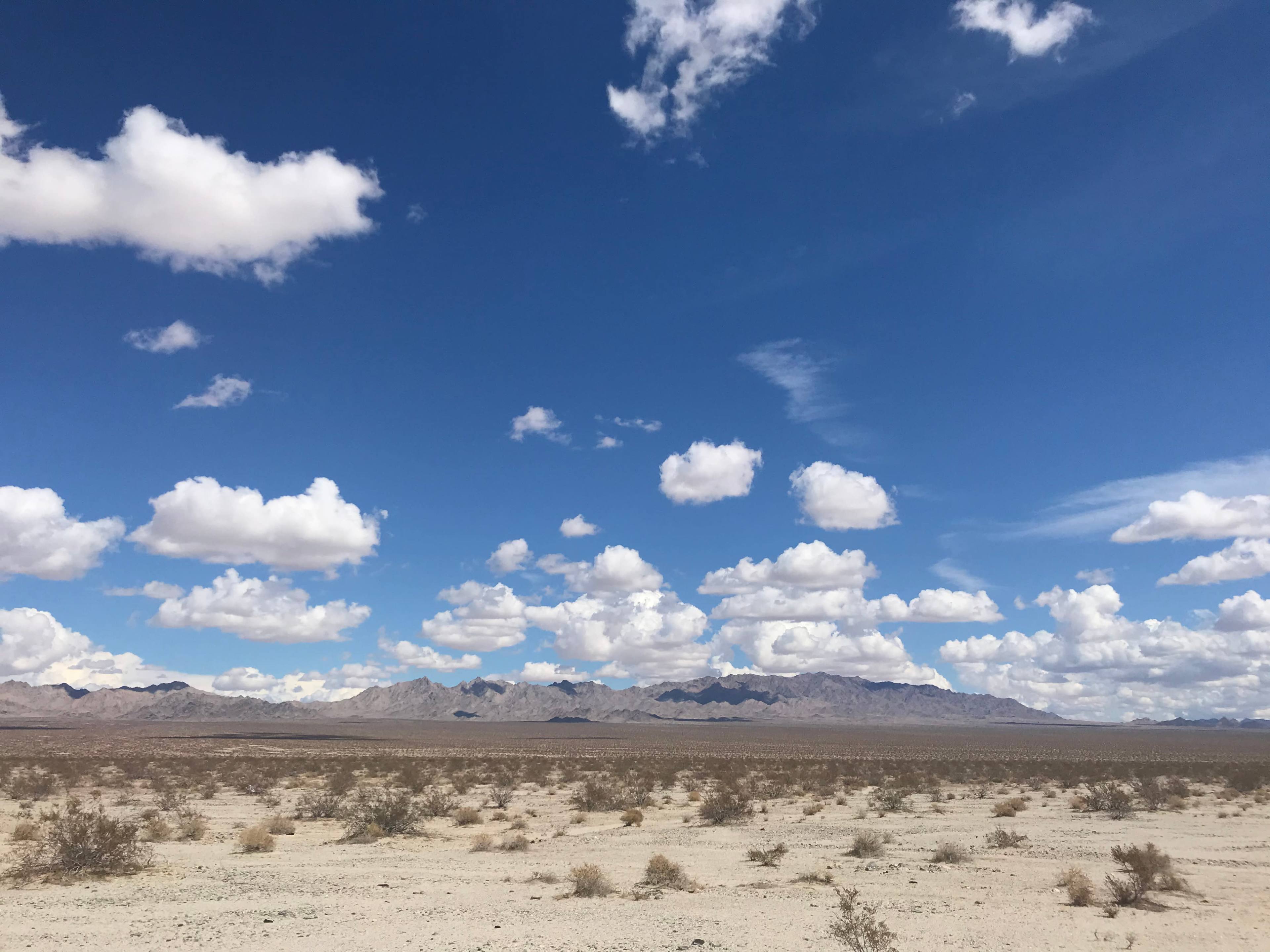 A vast desert landscape under a blue sky filled with scattered clouds and distant mountains.
