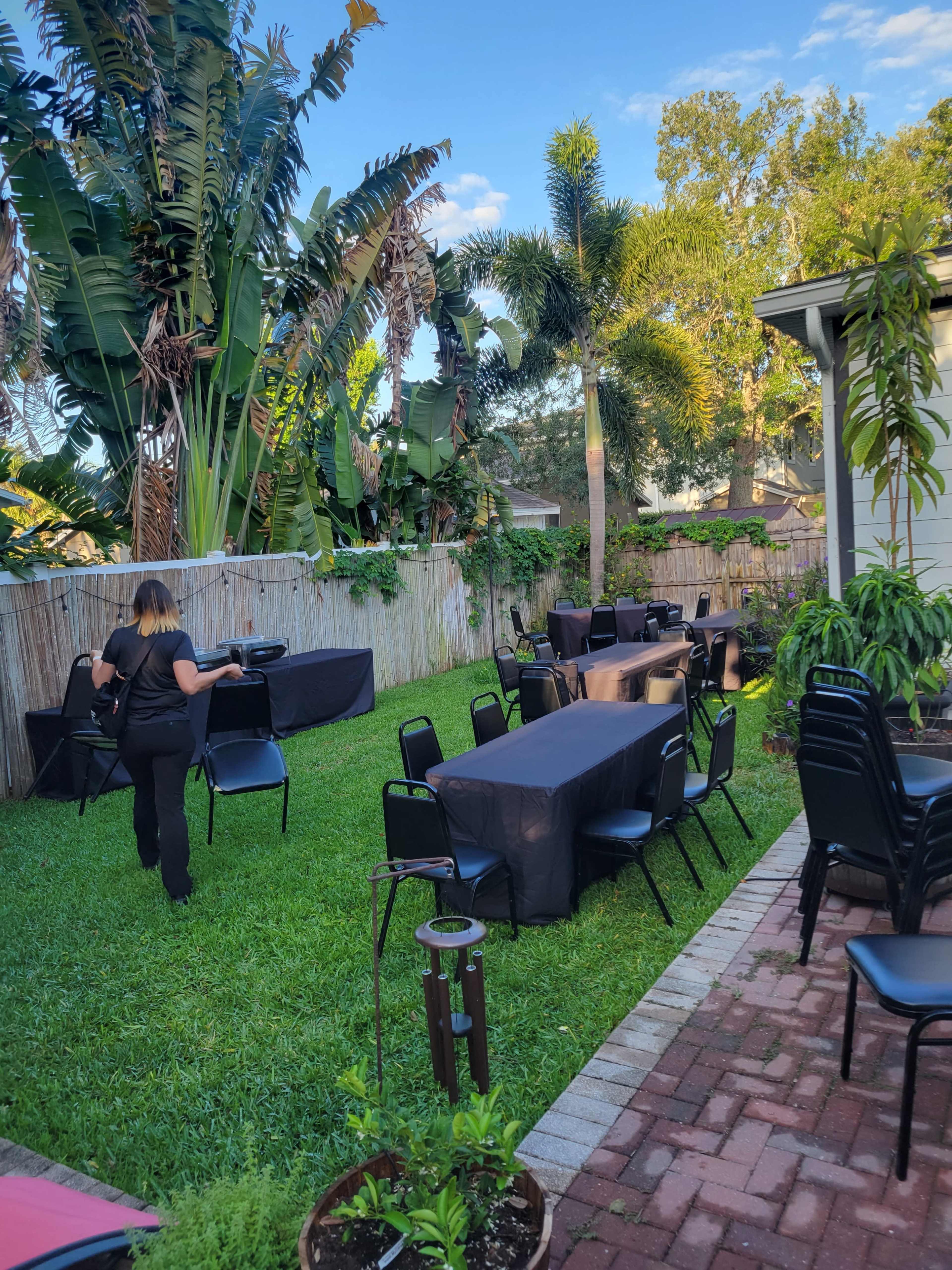 A person sets up black tables and chairs in a grassy backyard surrounded by tropical plants and trees.