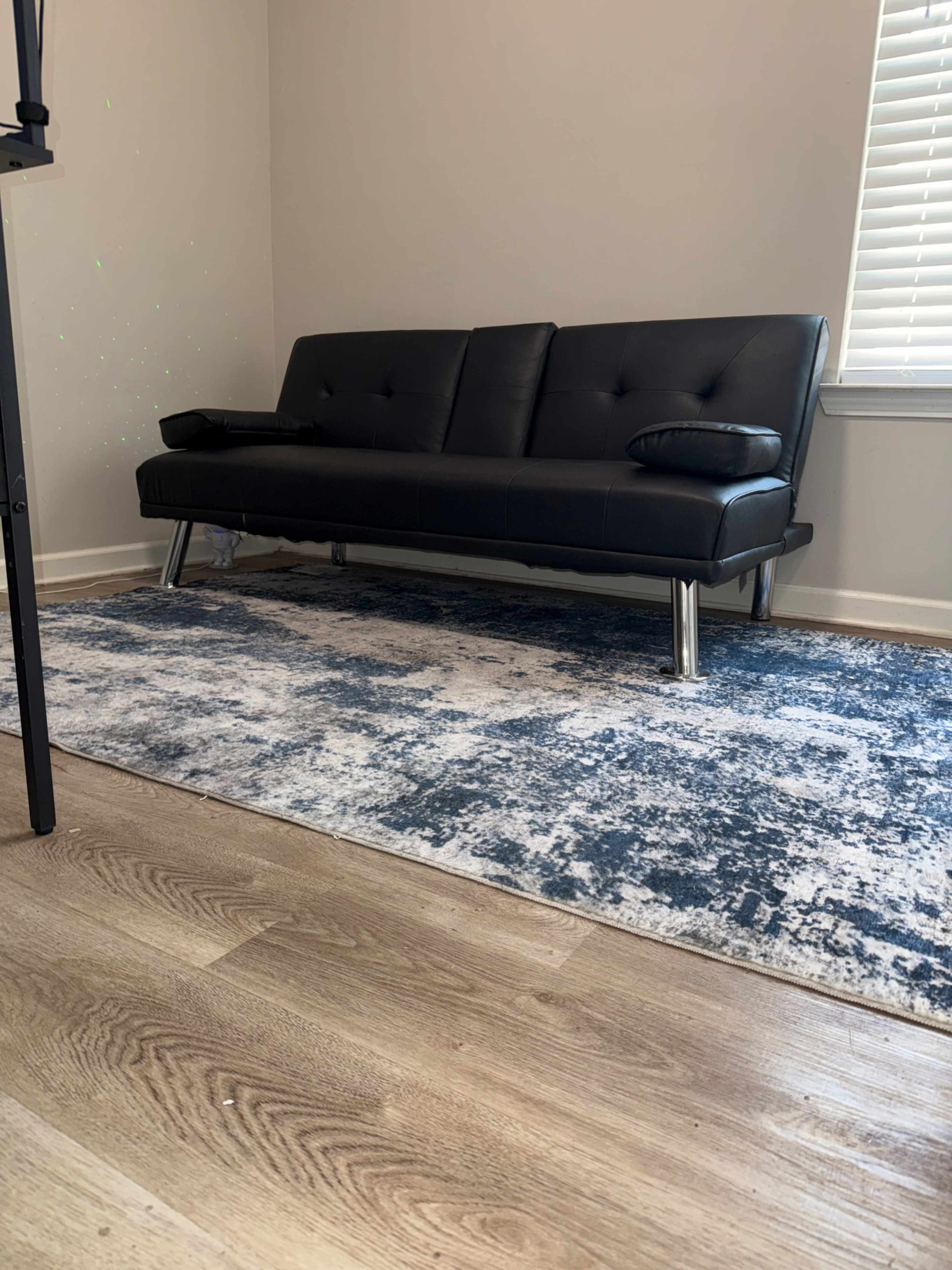 A black futon sits on a patterned blue and white area rug in a room with light-colored walls and a window.