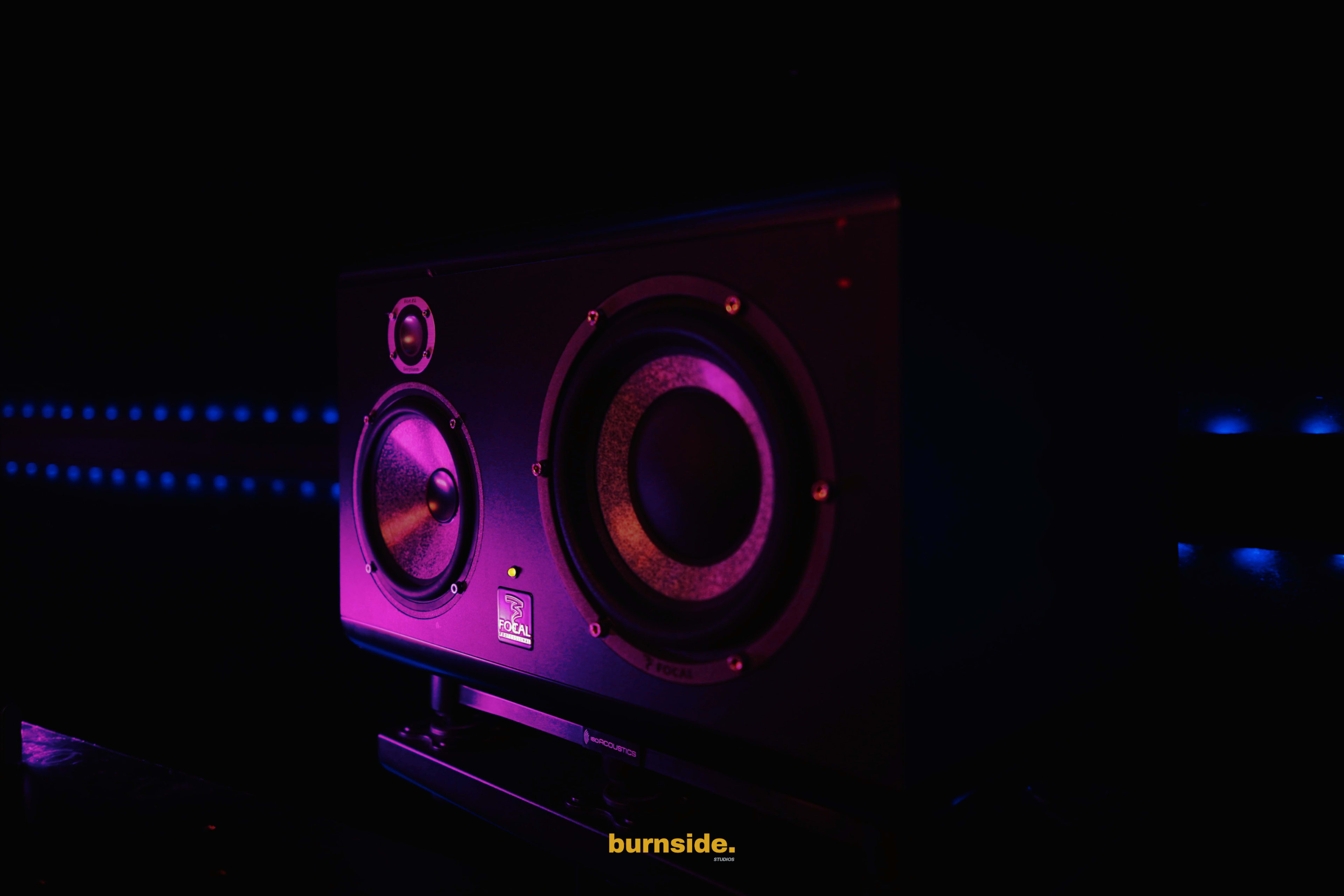 A black studio monitor speaker is illuminated with purple and blue light against a dark background.