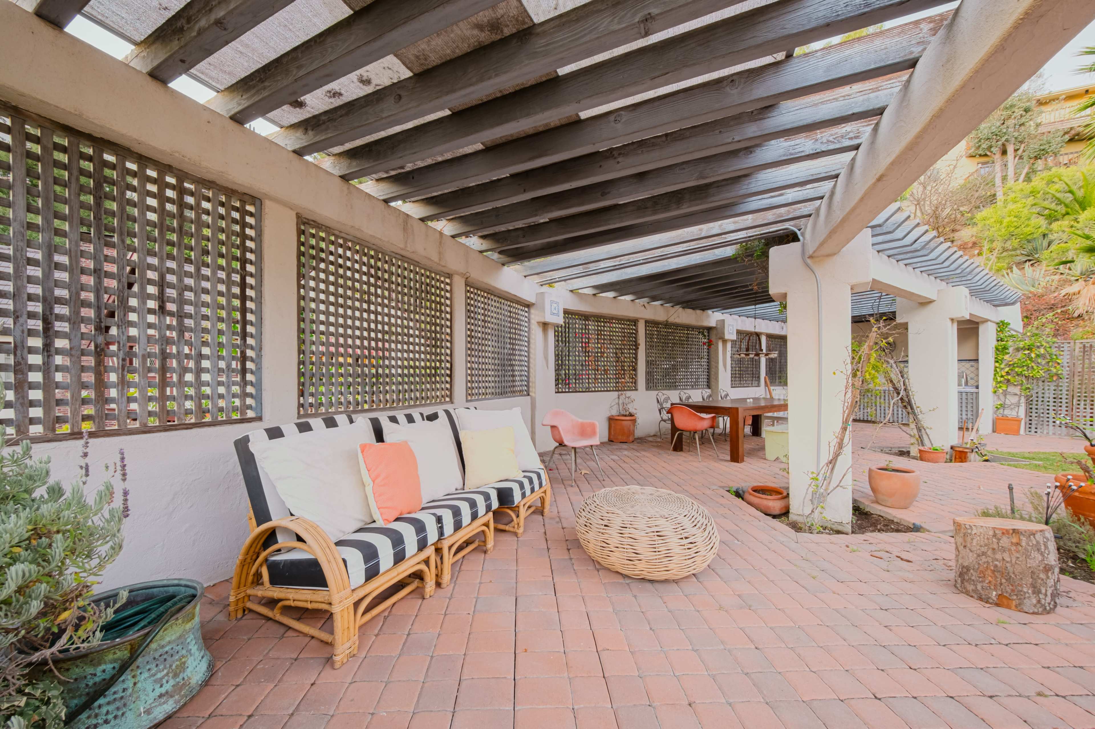 The image shows a covered patio area with a striped sofa, a round wicker coffee table, and potted plants.