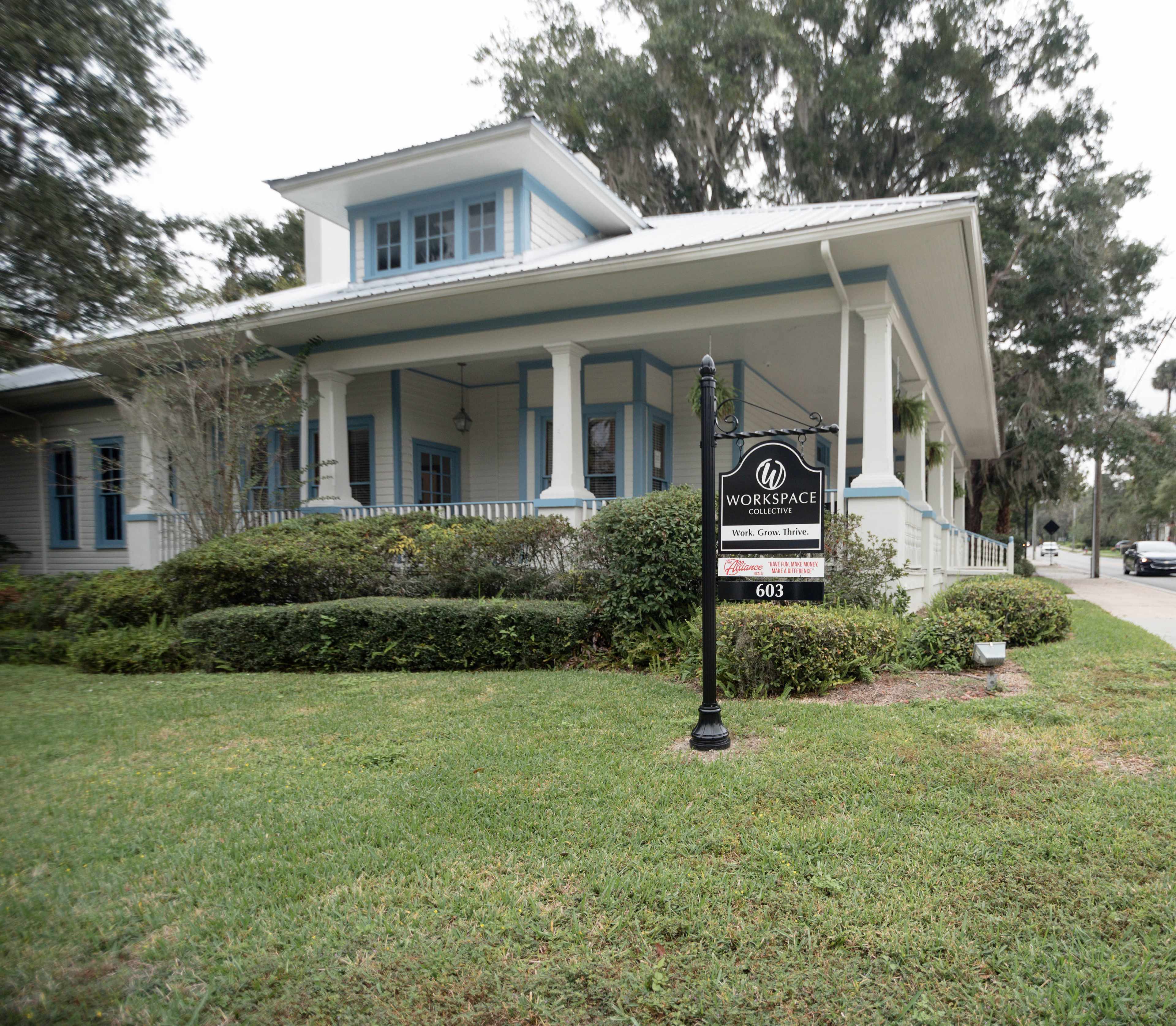 The image shows a large, light-colored building with a porch and a sign that reads "WORKSPACE" in front of a well-manicured lawn.