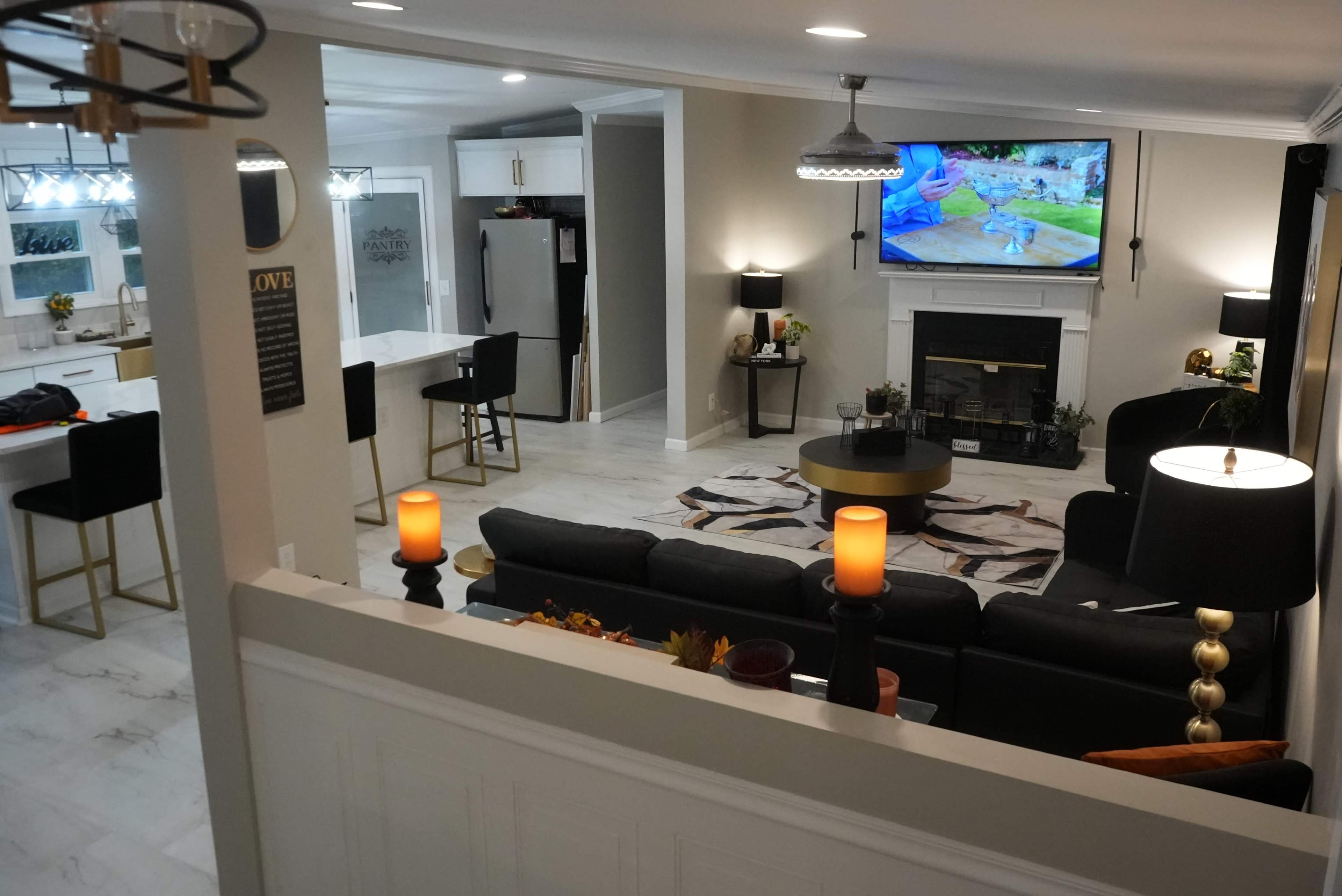 The image shows a modern living room with a black sofa, a round coffee table, and a television mounted above a fireplace, all set against a backdrop of light-colored walls and floors.