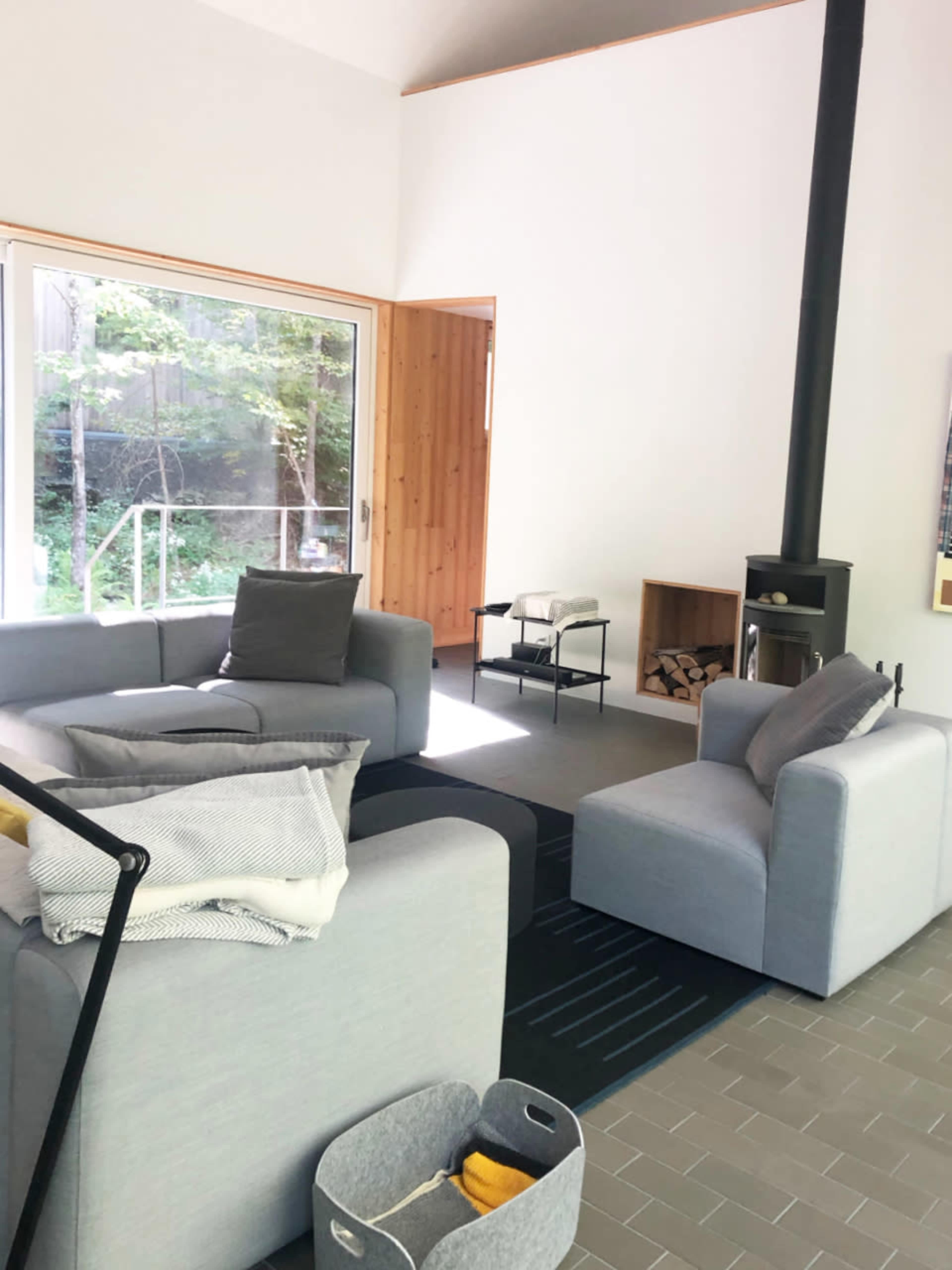 A modern living room features gray sofas, a black wood stove, and large windows overlooking greenery.