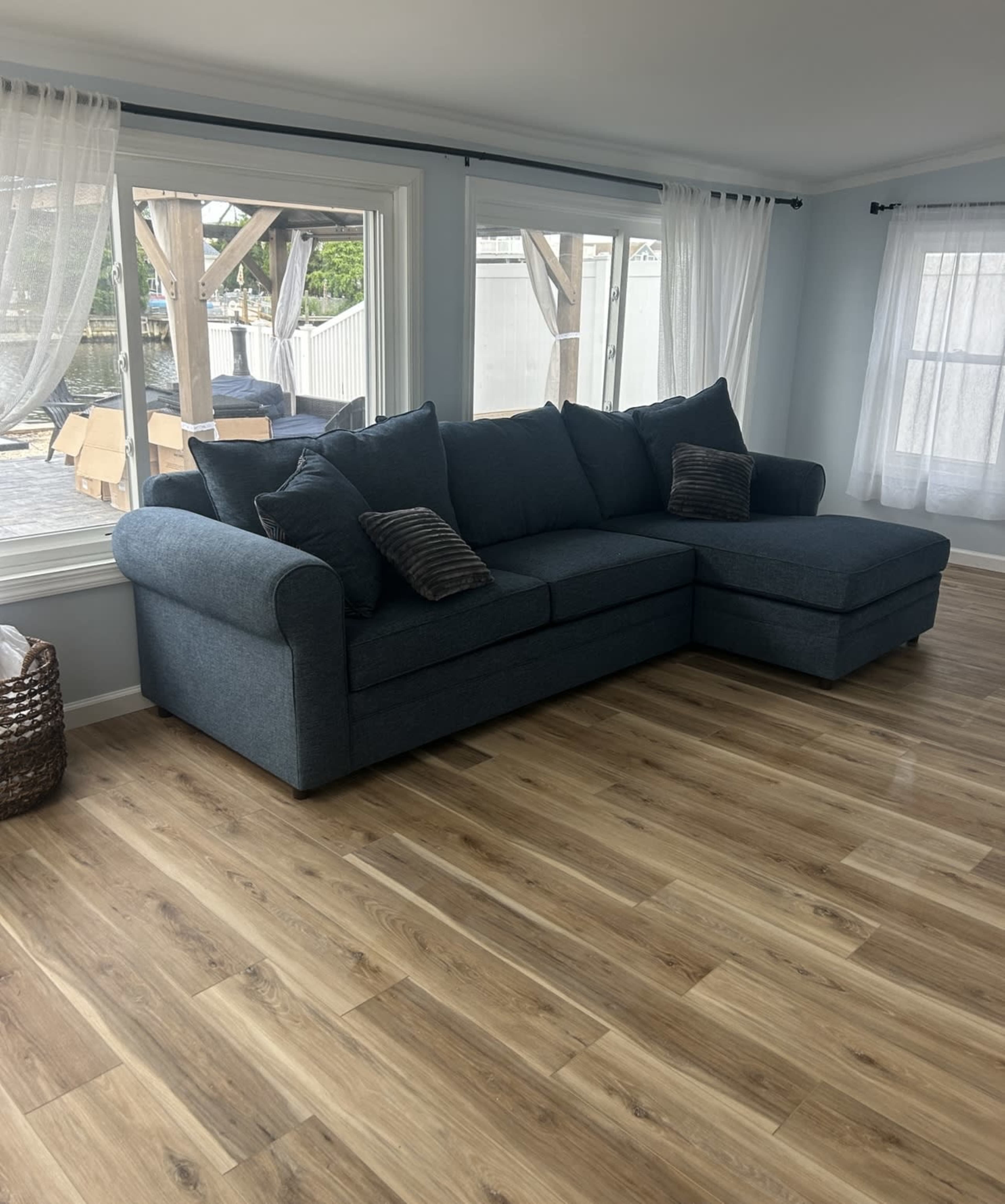 A blue sectional sofa is positioned in a bright room with large windows and light wood flooring.