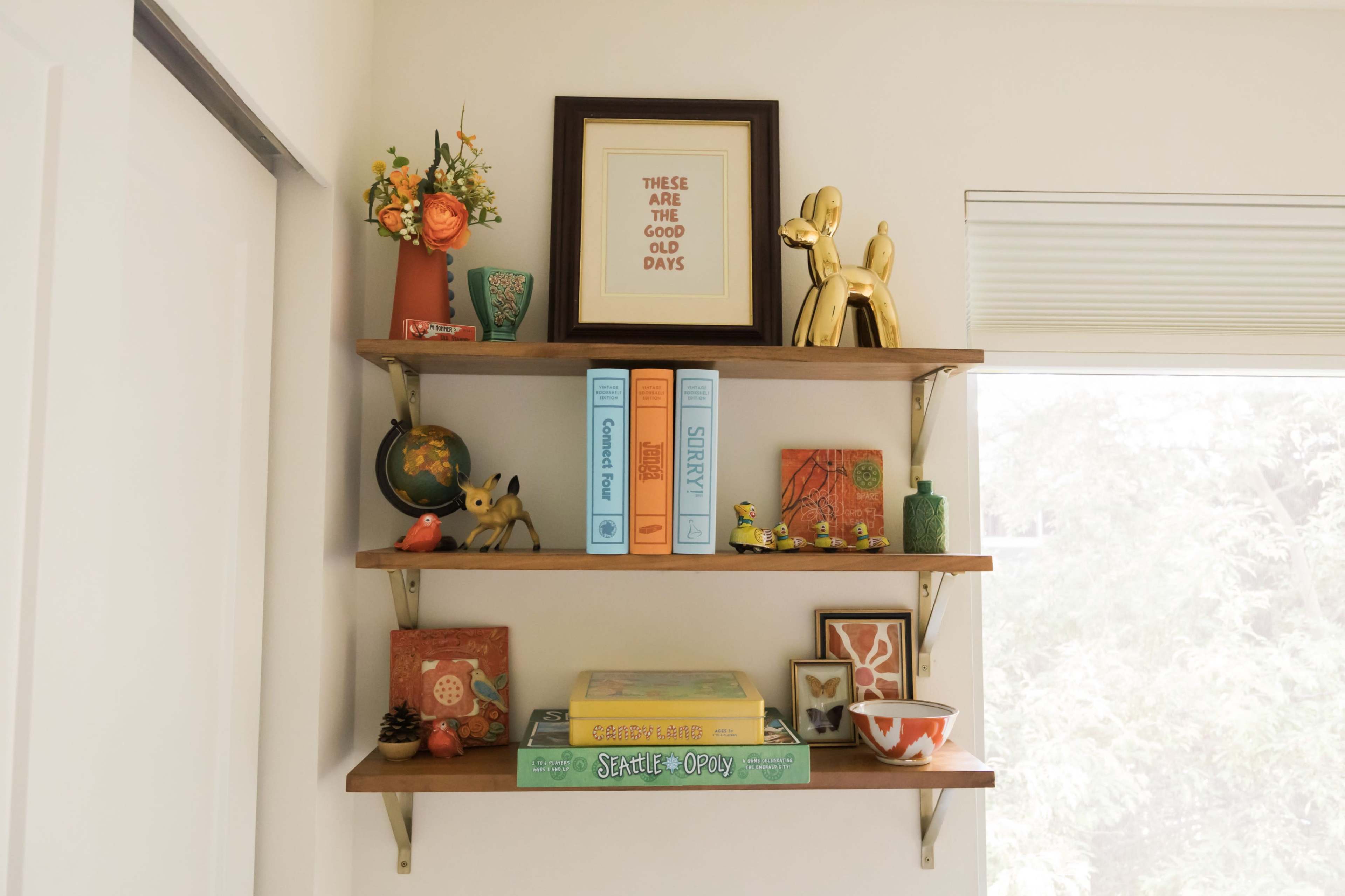 The image features a set of three wooden shelves displaying a collection of decorative items, books, and framed art in a well-lit room.