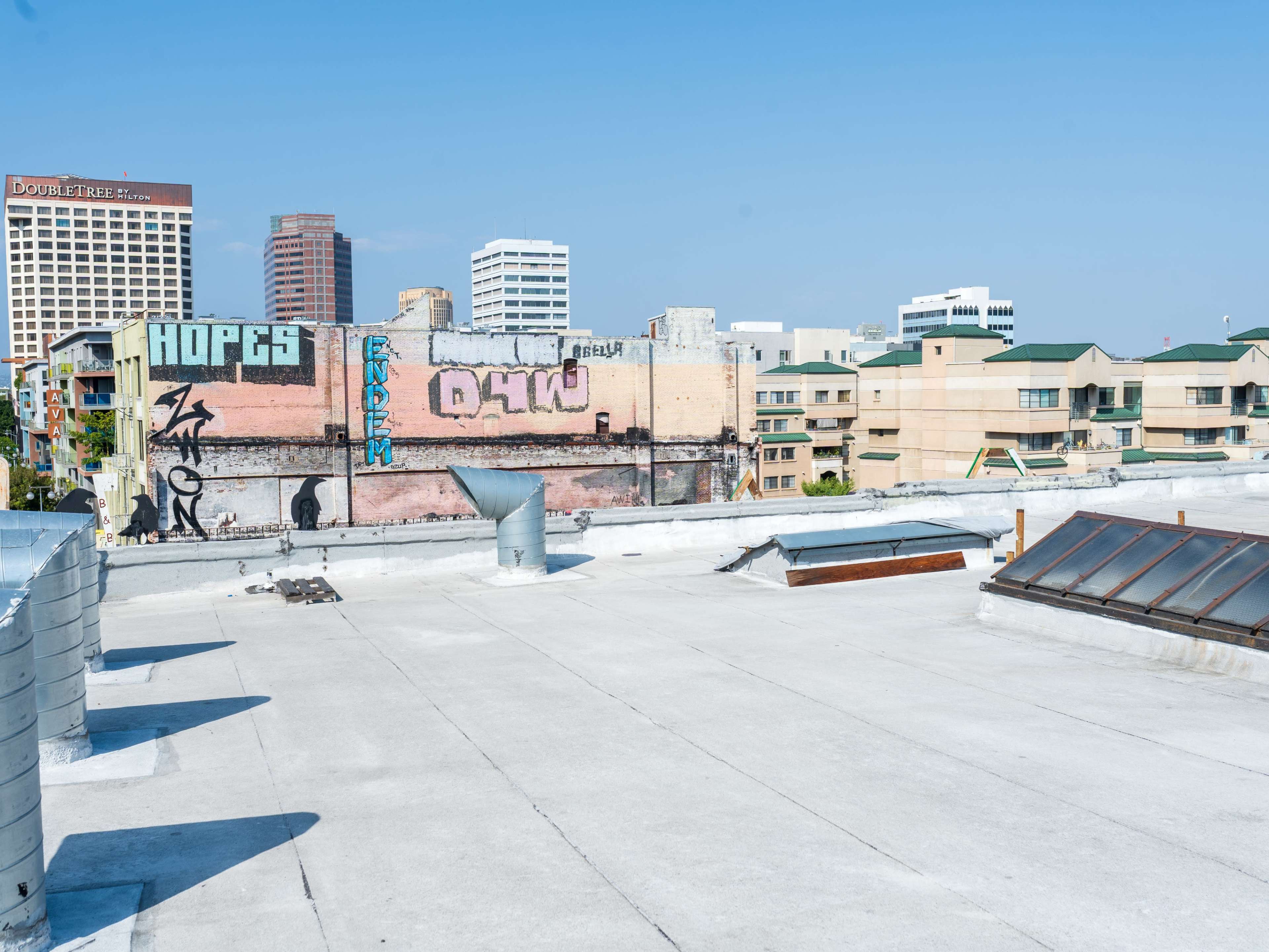 The image shows a flat rooftop with industrial ventilation systems and graffiti-covered walls set against a skyline of buildings in an urban environment.