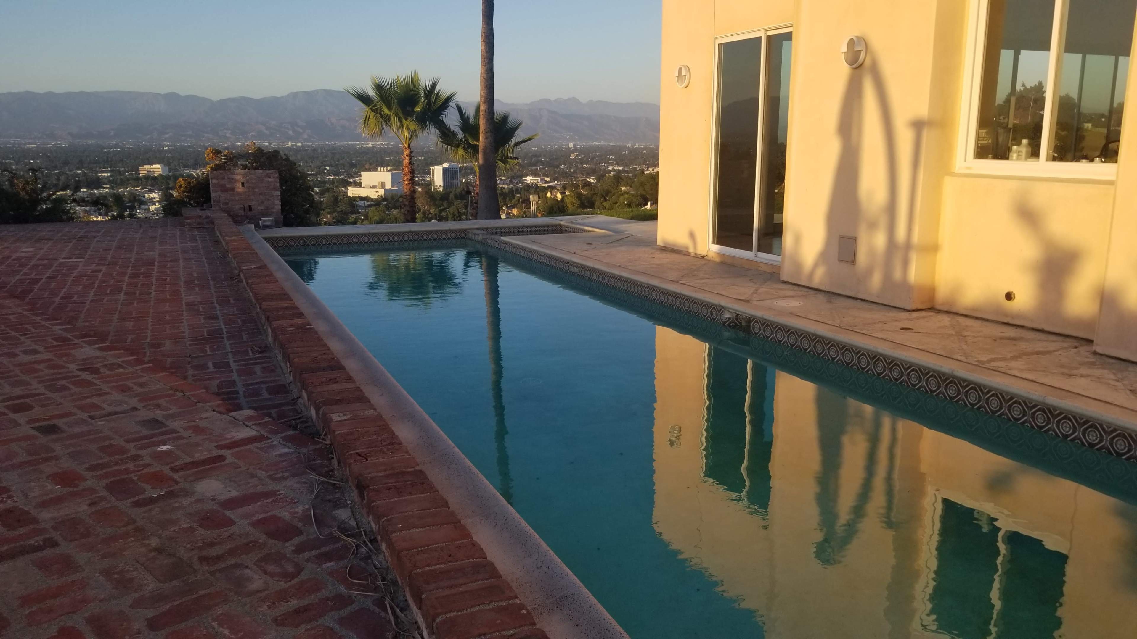 A clear swimming pool reflecting a building and mountains, with a brick patio in the foreground.