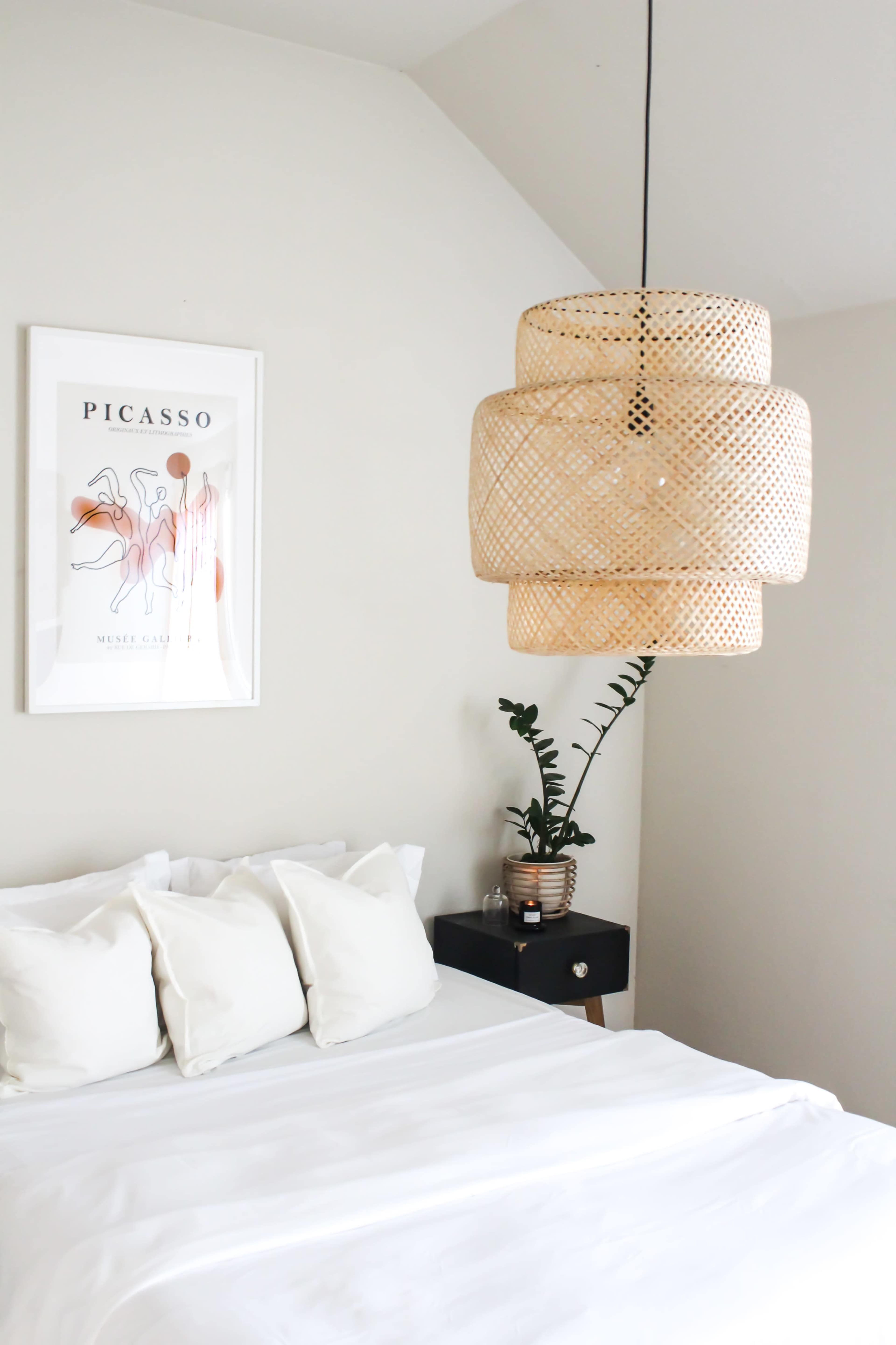 A bedroom features a large woven pendant light above a neatly made bed with white pillows, alongside a plant and framed art on the wall.