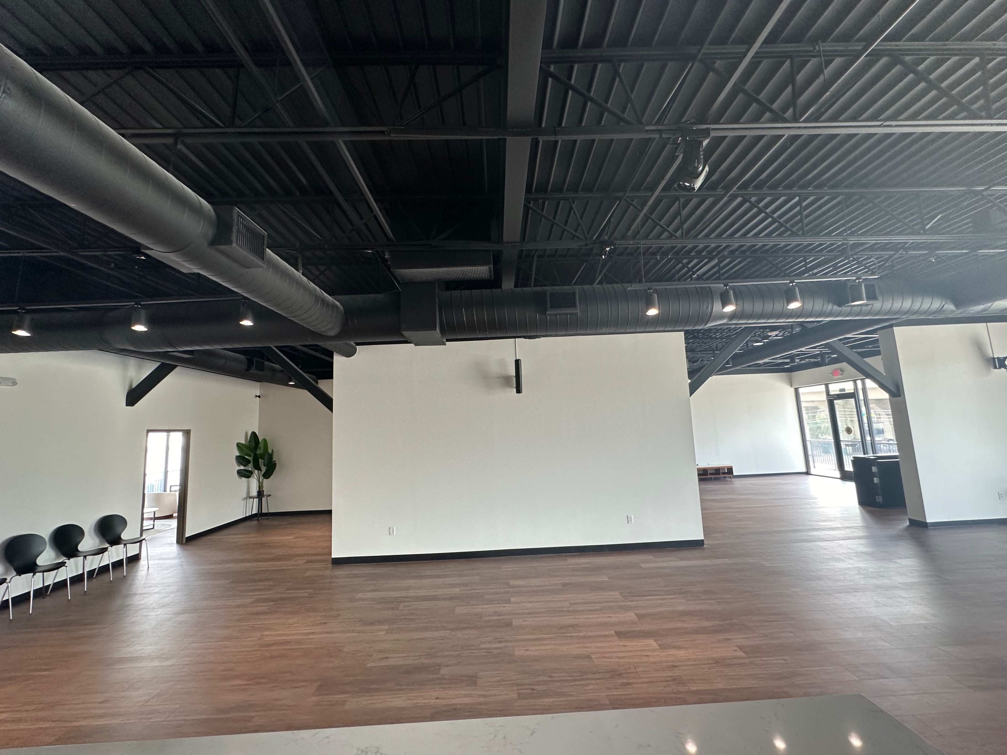 The image shows a spacious, modern interior with dark beams, hardwood flooring, and minimal furniture.