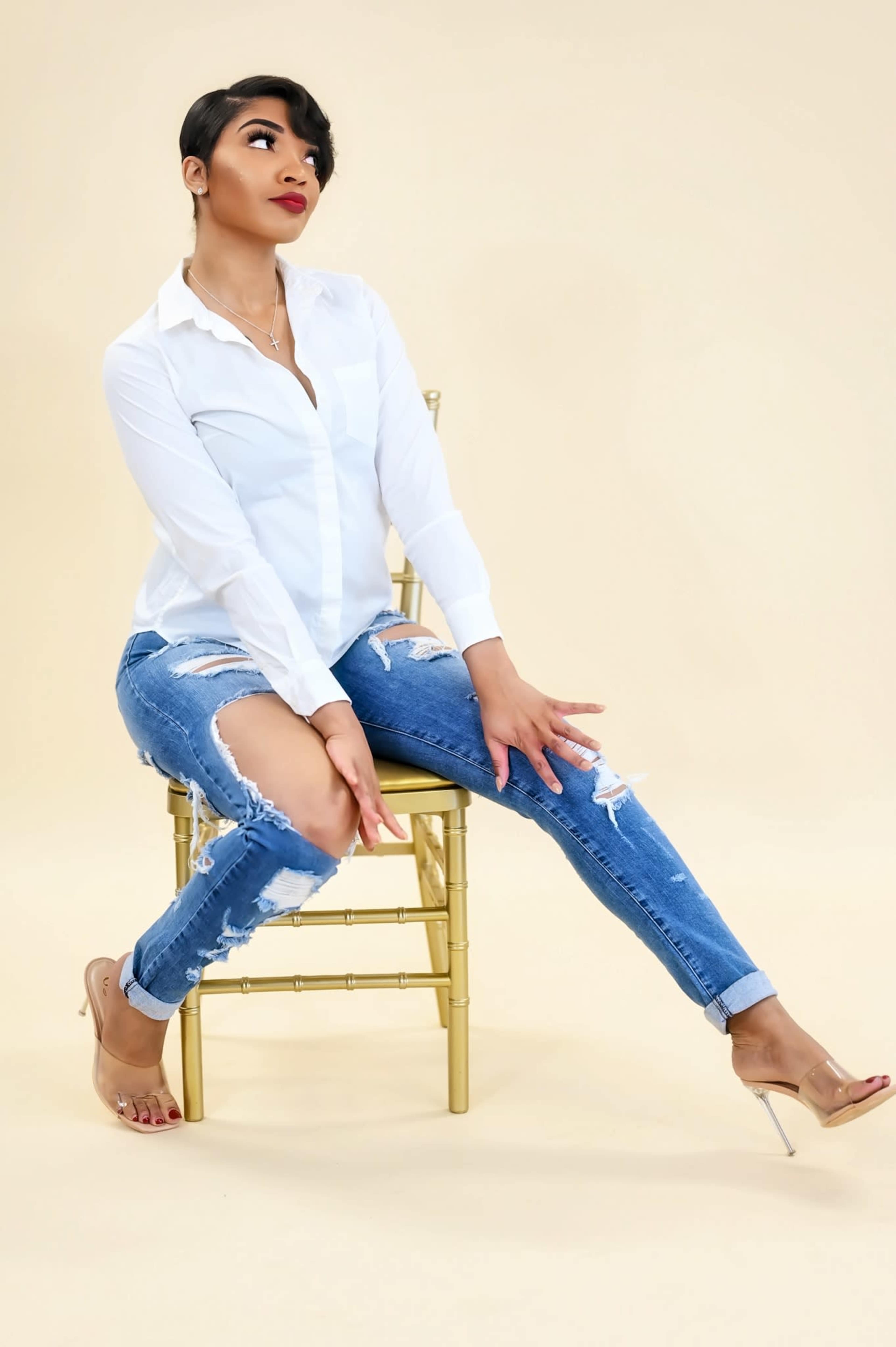 A woman in a white blouse and distressed jeans sits on a gold chair against a light beige background.