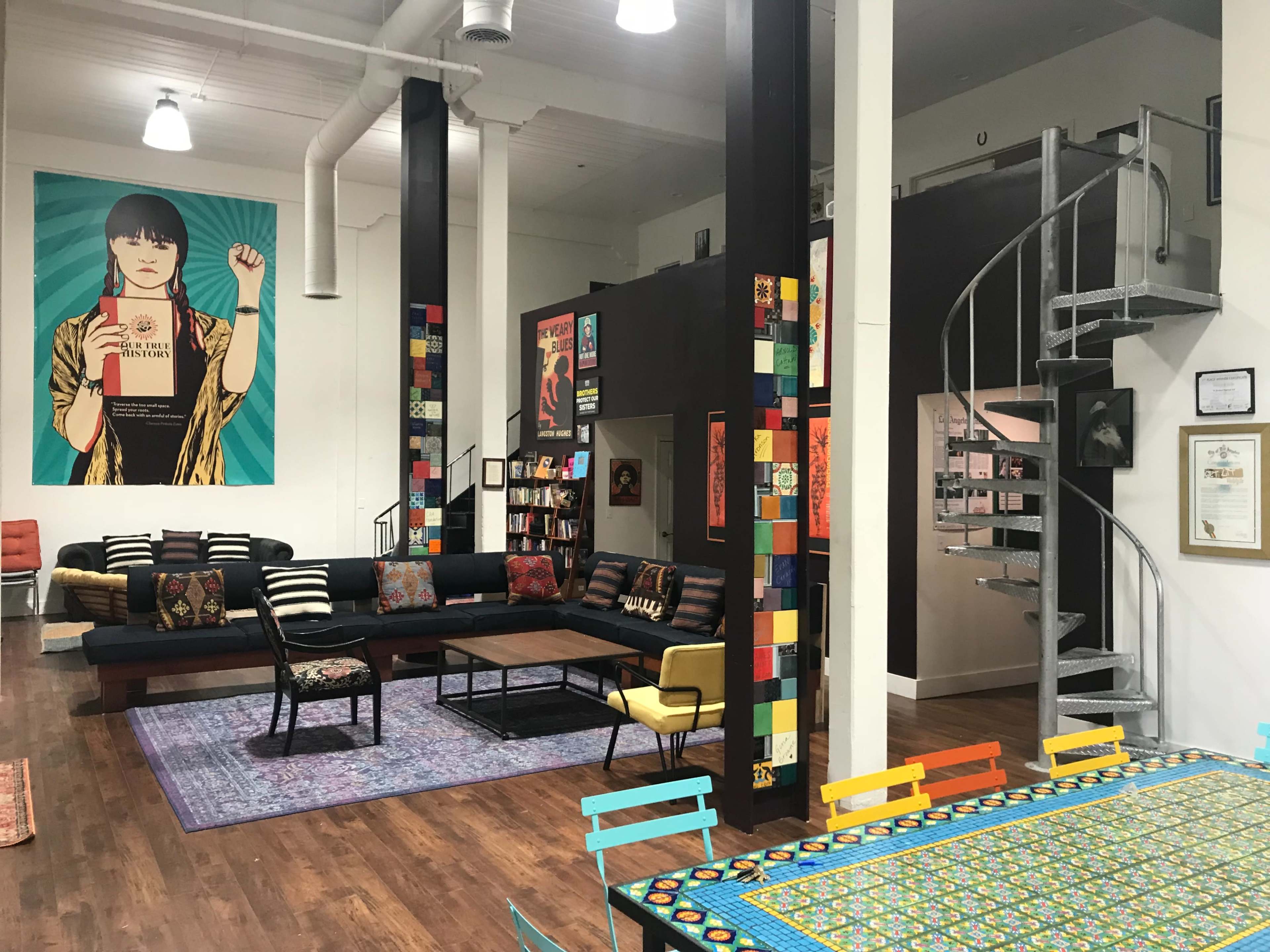 The image shows a stylishly decorated open space featuring a spiral staircase, a colorful dining table, a black couch with patterned pillows, and large vibrant wall art.