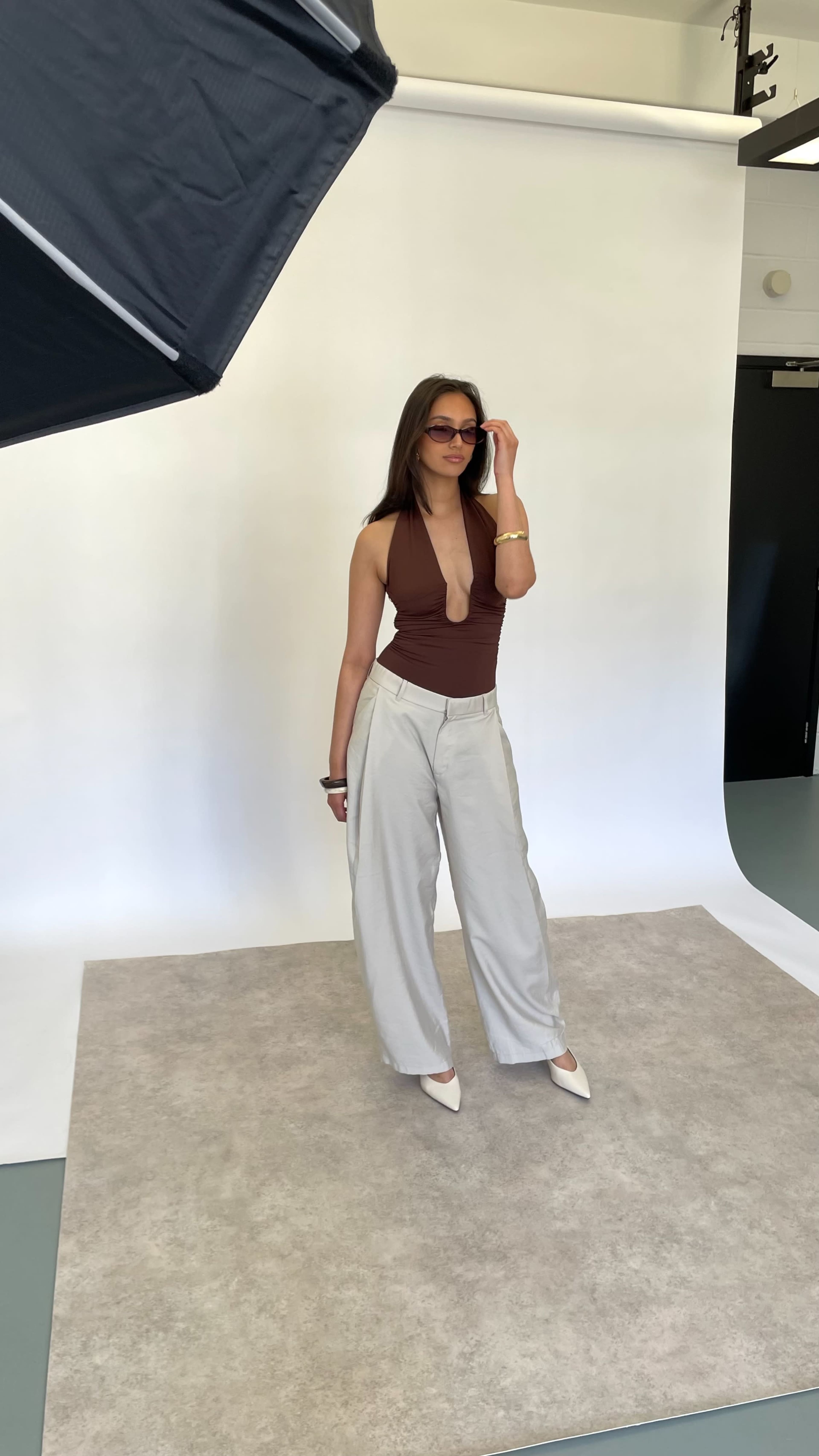 A model poses on a neutral backdrop, wearing a brown top, light gray wide-legged pants, and sunglasses.