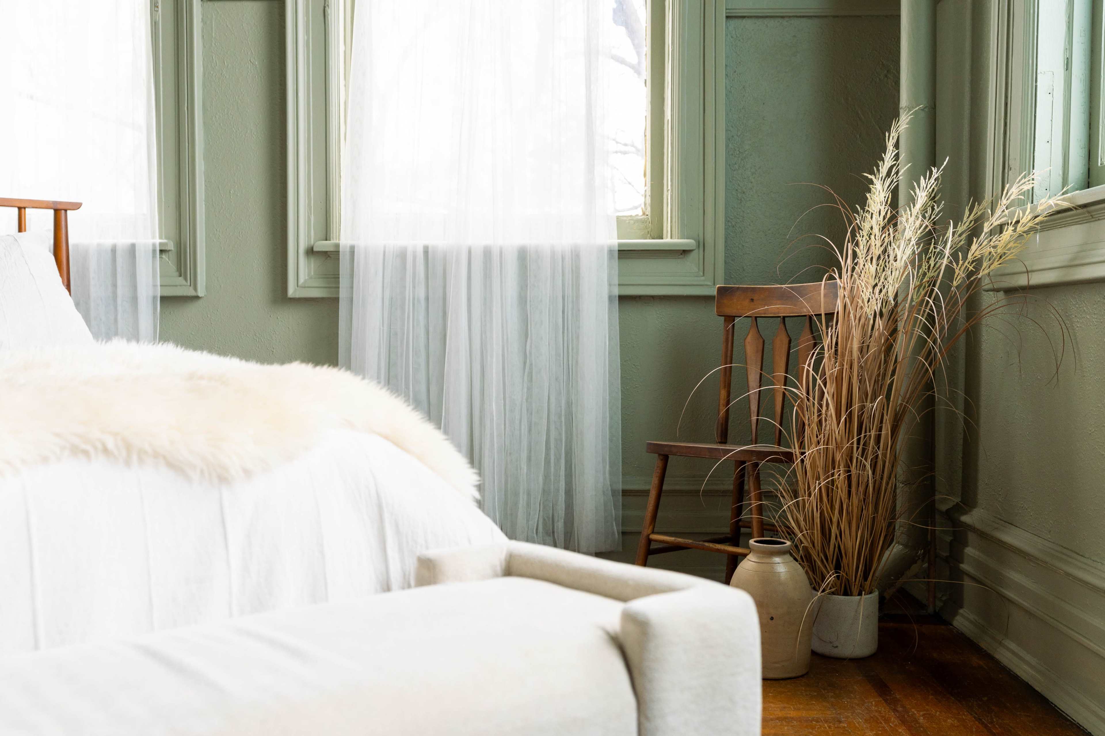The image shows a softly lit bedroom featuring a bed with a light-colored blanket, a wooden chair beside a vase with dried plants, and sheer curtains that allow natural light to enter.