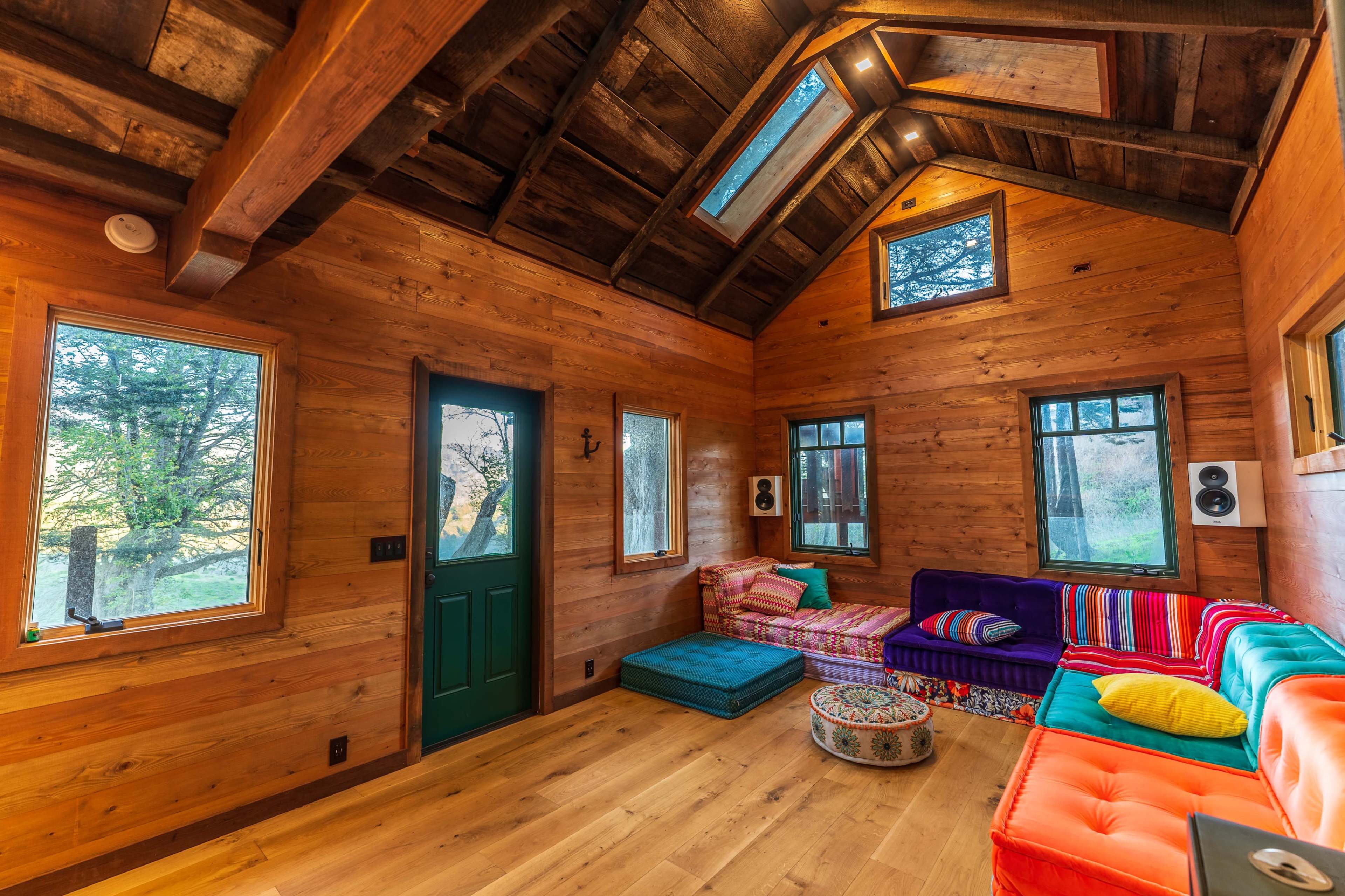 A small, wooden interior features a variety of colorful couches and large windows that allow natural light to enter.