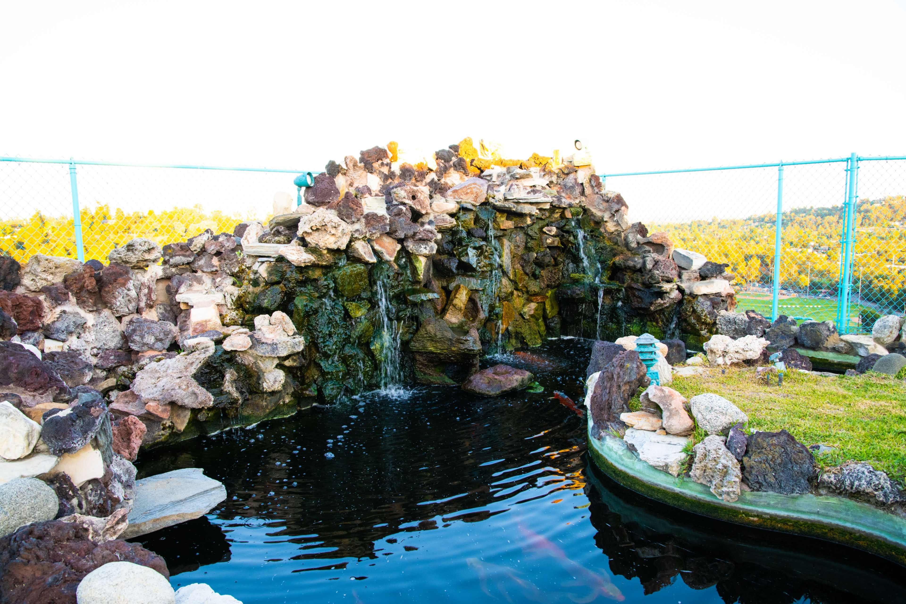 A stone water feature with cascading water sits beside a small pond, surrounded by a fenced grassy area.