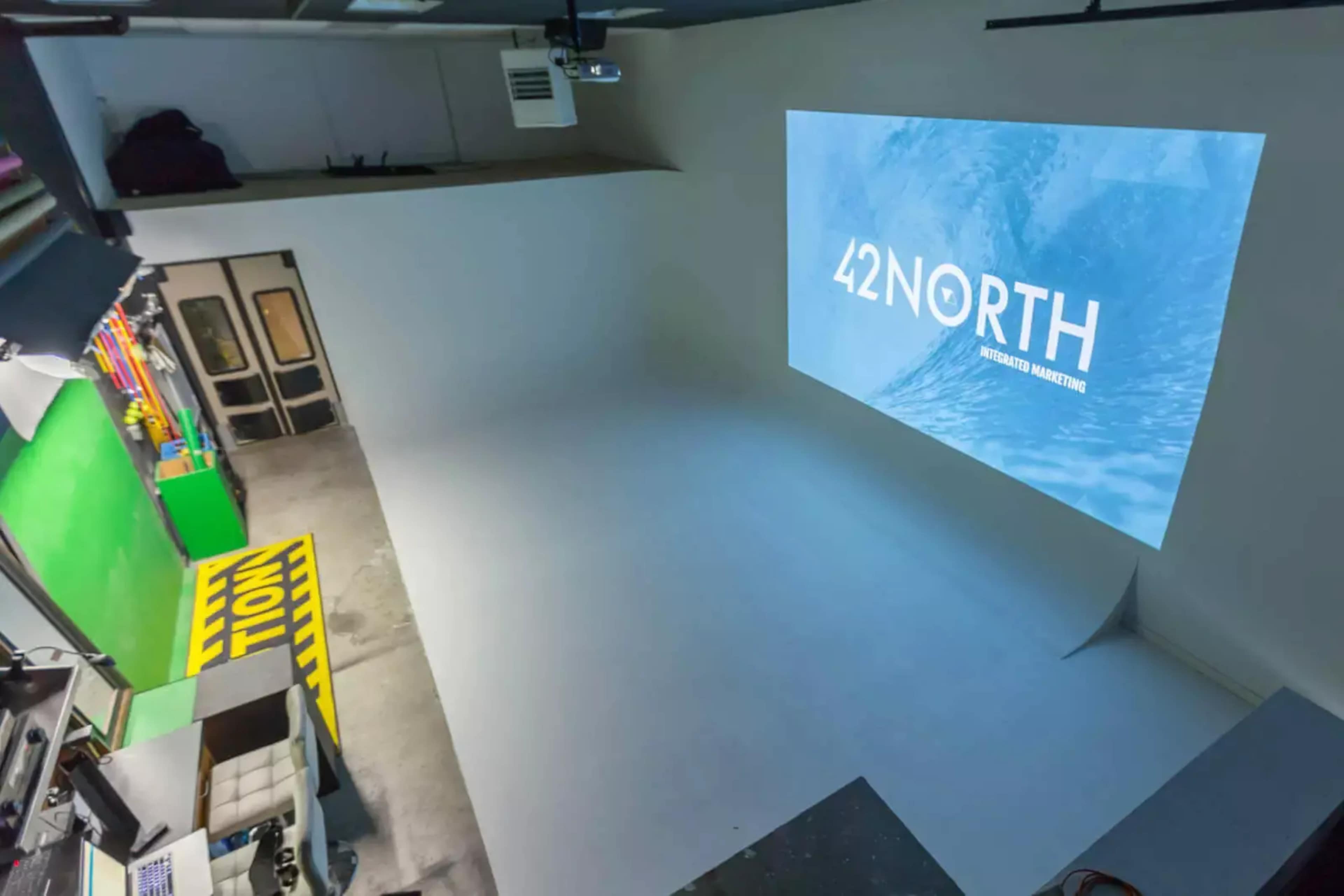 The image shows a spacious studio featuring a blank white backdrop and a large projected screen displaying "42 NORTH."