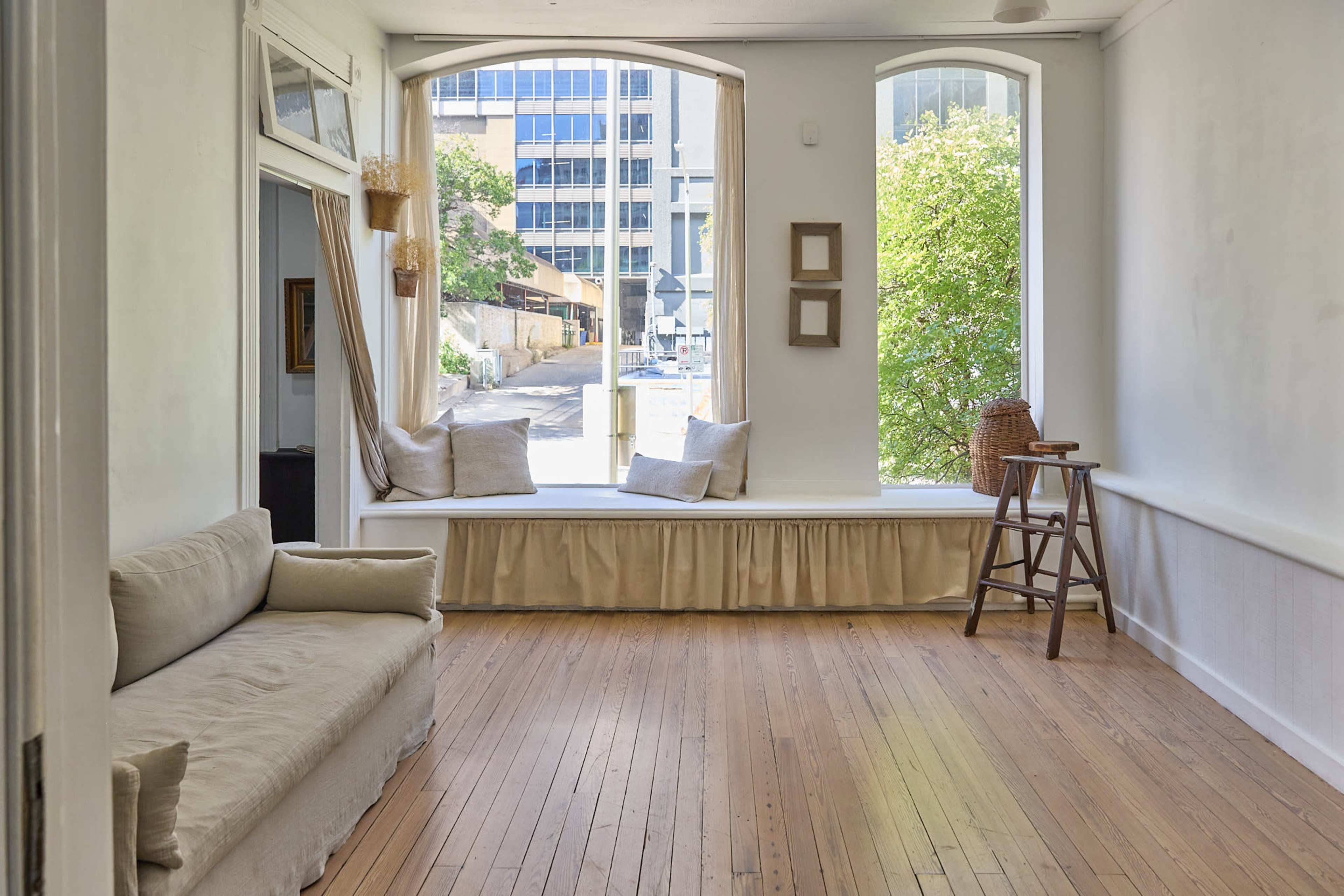 The image shows a minimalist interior of a room with wooden flooring, featuring a beige sofa, a window seat with cushions, and a small wooden stool beside it.
