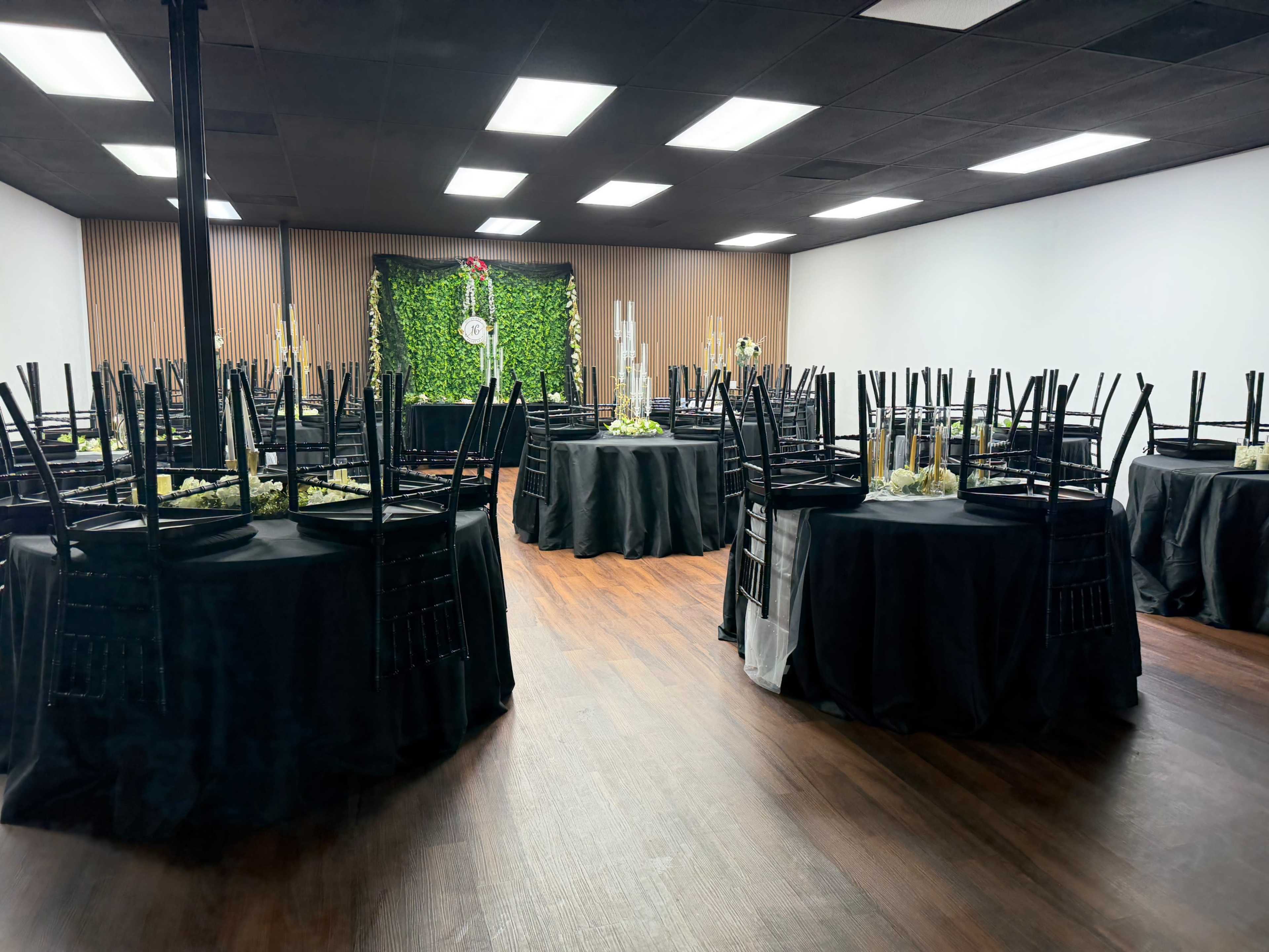 A banquet hall is arranged with black tablecloths and stacked chairs, featuring a green backdrop decorated with candles and floral arrangements.