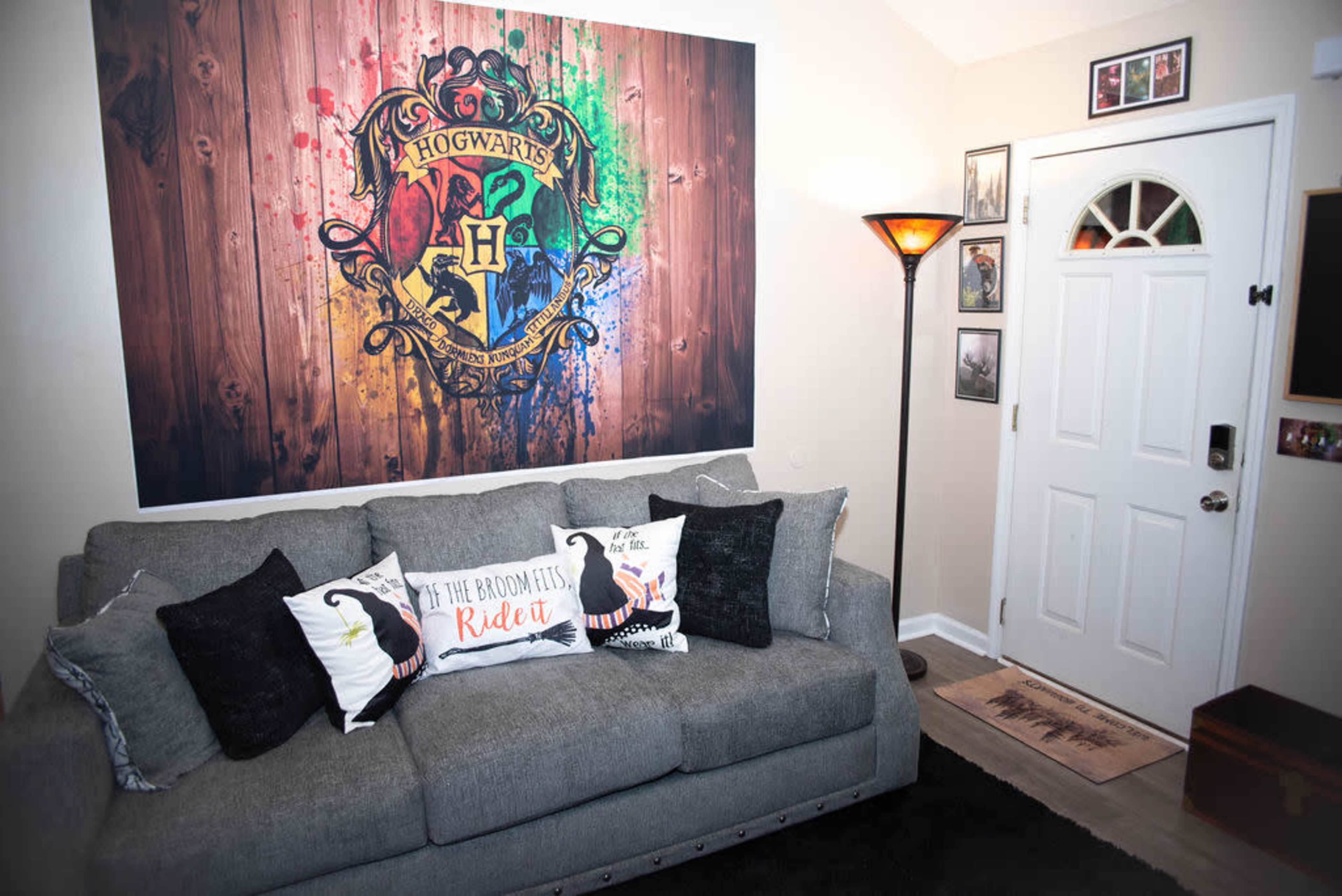 A cozy living room featuring a grey couch with decorative pillows, a large Hogwarts crest mural on the wall, and a front door with a lamp and framed photos nearby.