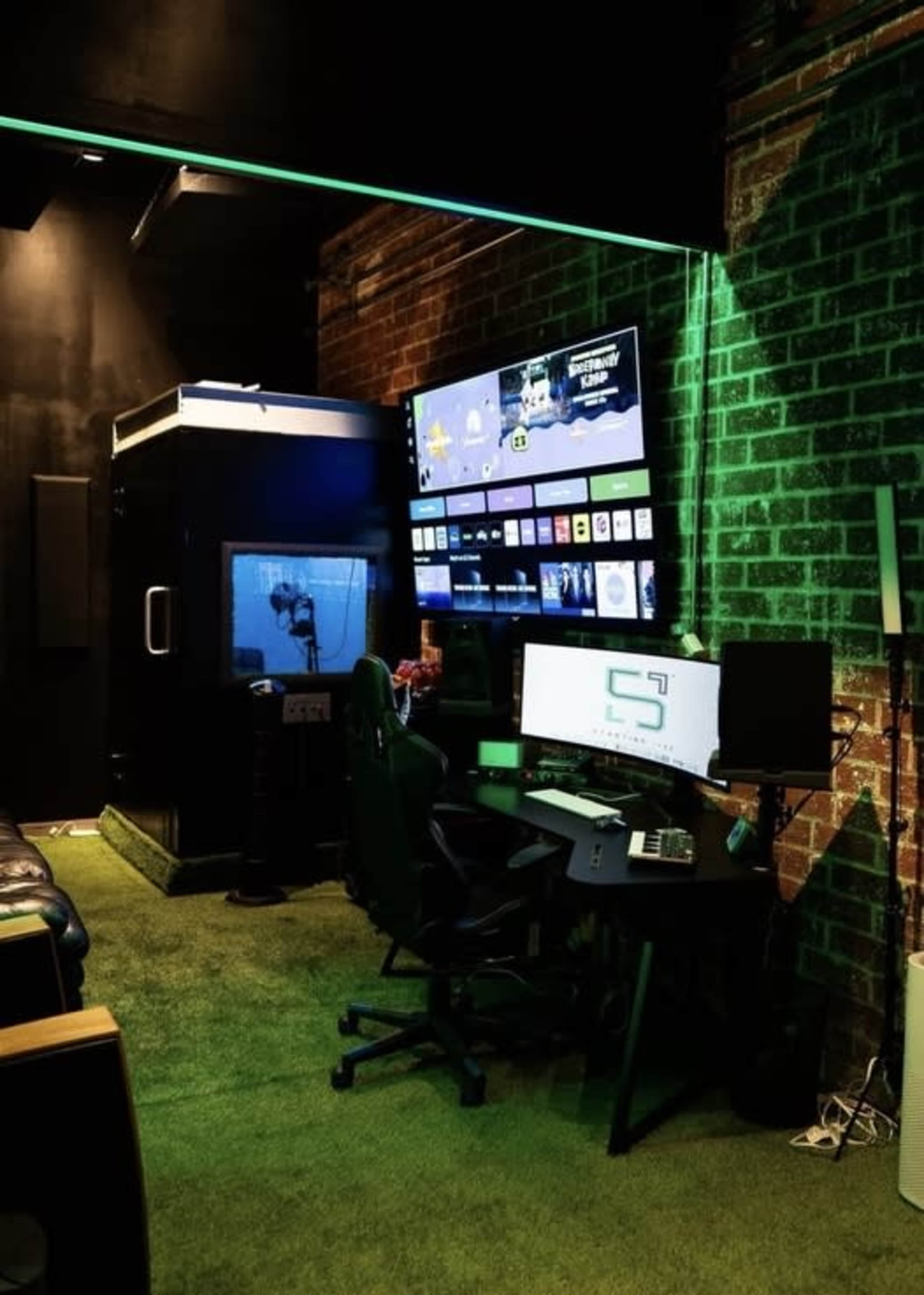 The image features a modern gaming setup with multiple screens, a black gaming chair, and illuminated green accents in a brick-walled room.