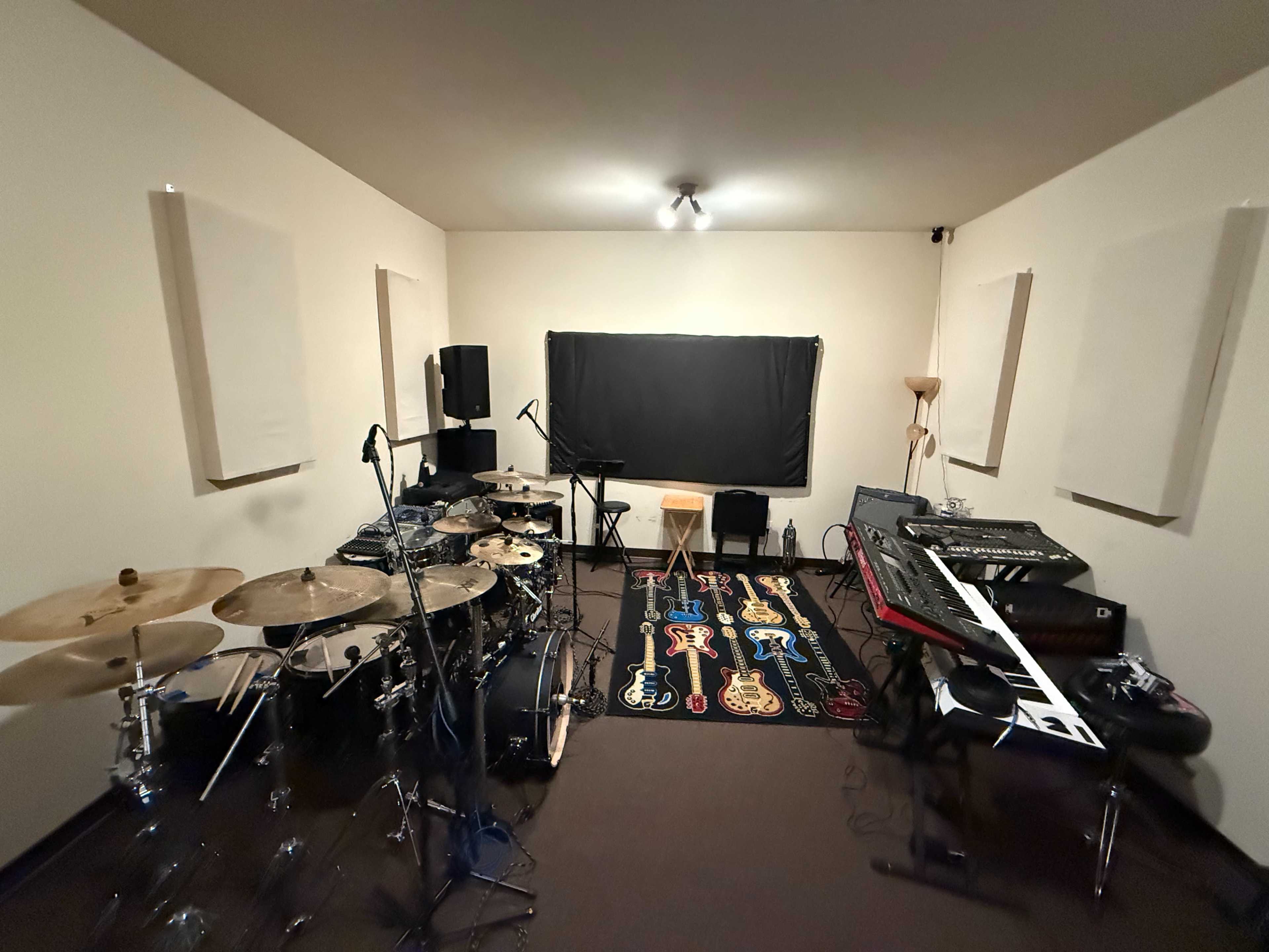 The image shows a music rehearsal room with a drum set, keyboards, and sound equipment arranged around the space.