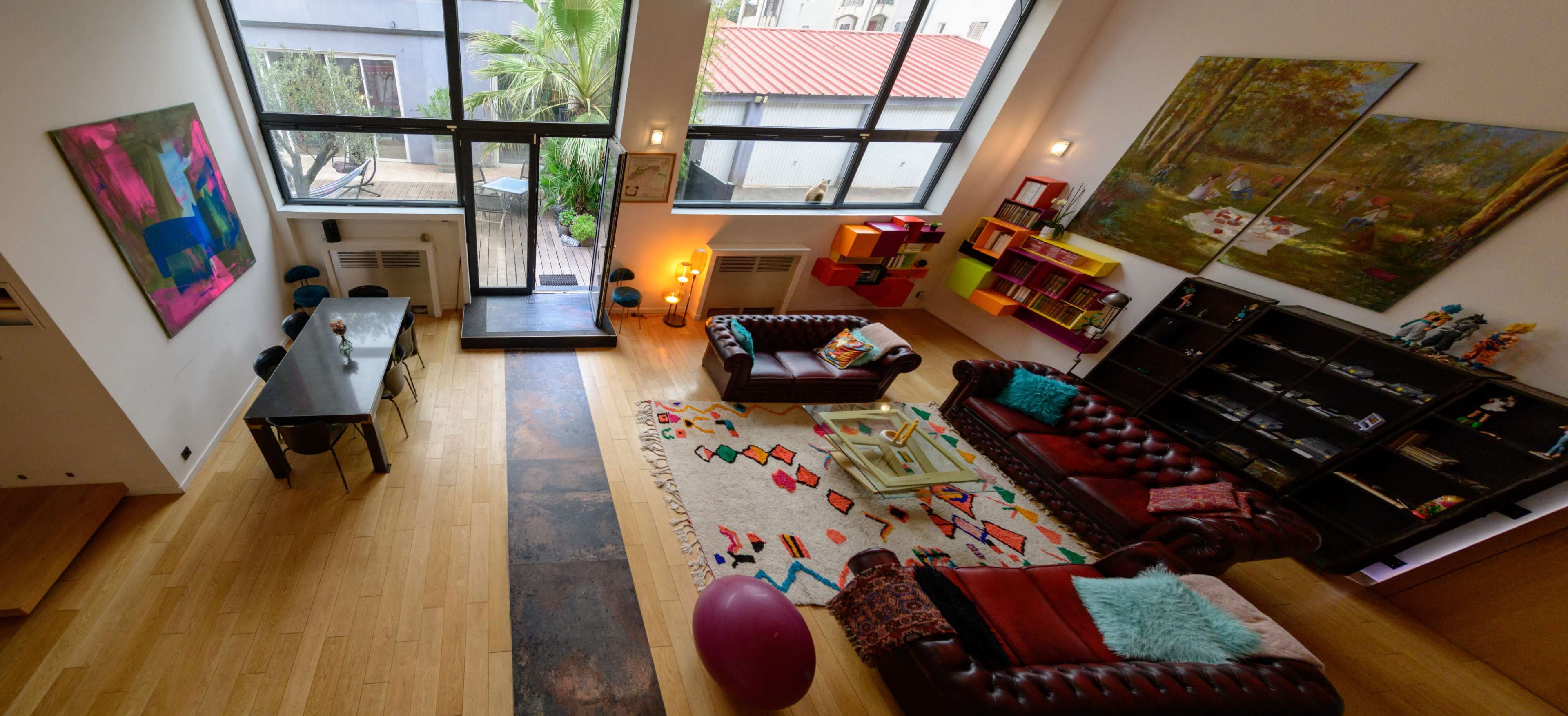 The image shows a spacious, modern living area with large windows, featuring a dining table, two leather sofas, colorful artwork, and a vibrant rug.