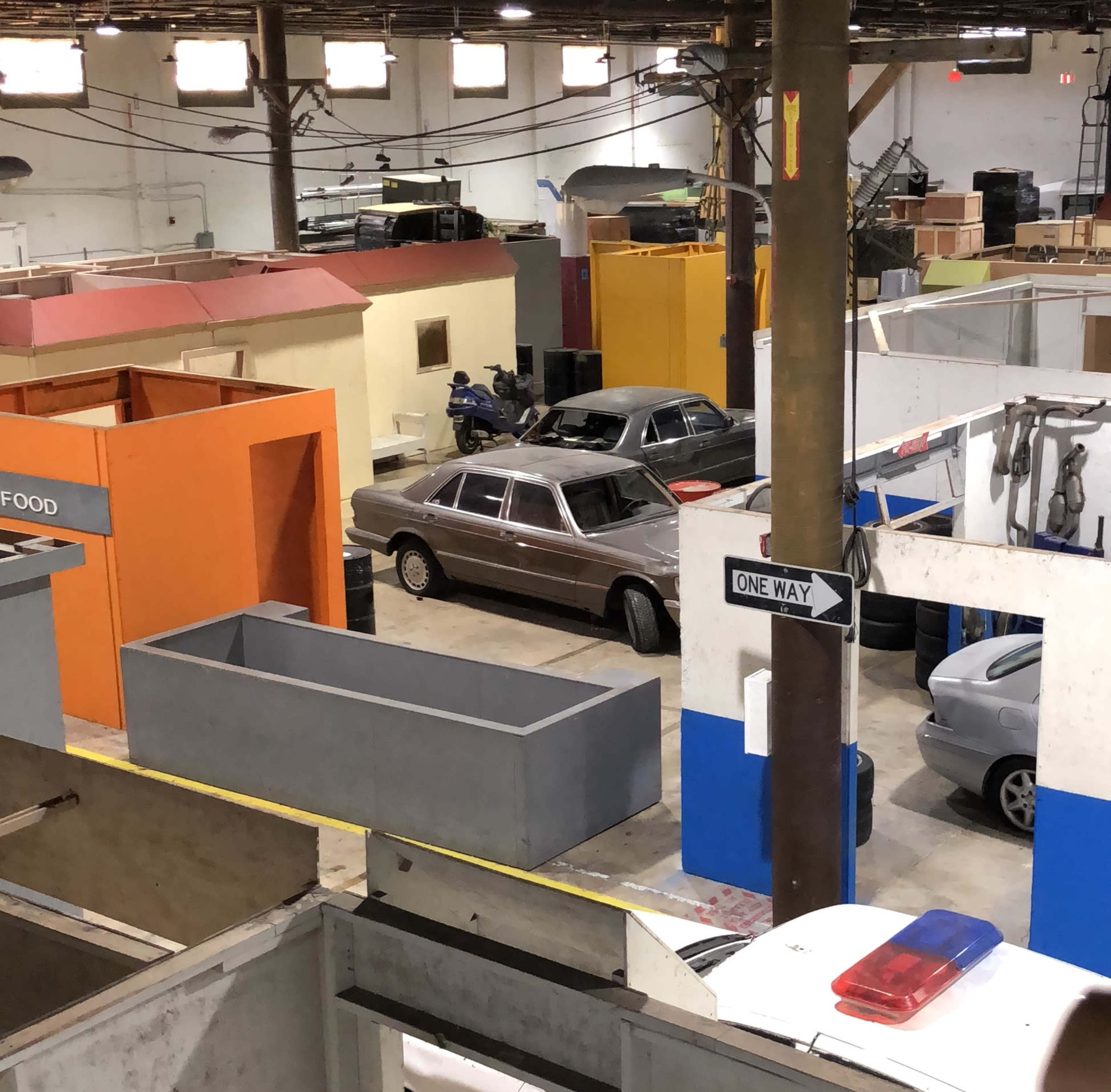 The image shows a large indoor space resembling a set or training area, featuring multiple colored structures and two parked cars.