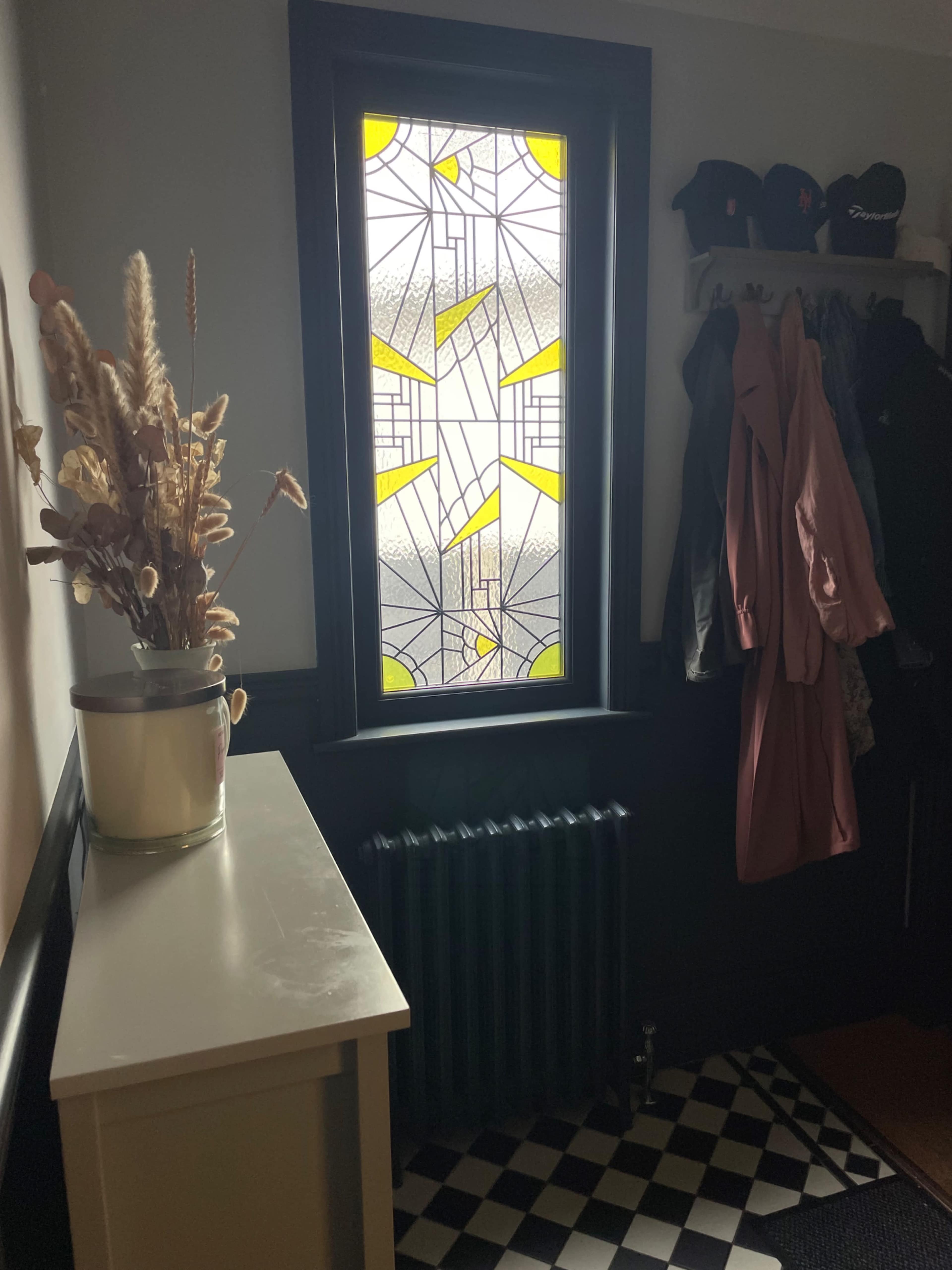 The image shows a hallway with a stained glass window, a small table with a vase of dried flowers, and hanging coats next to a radiator.