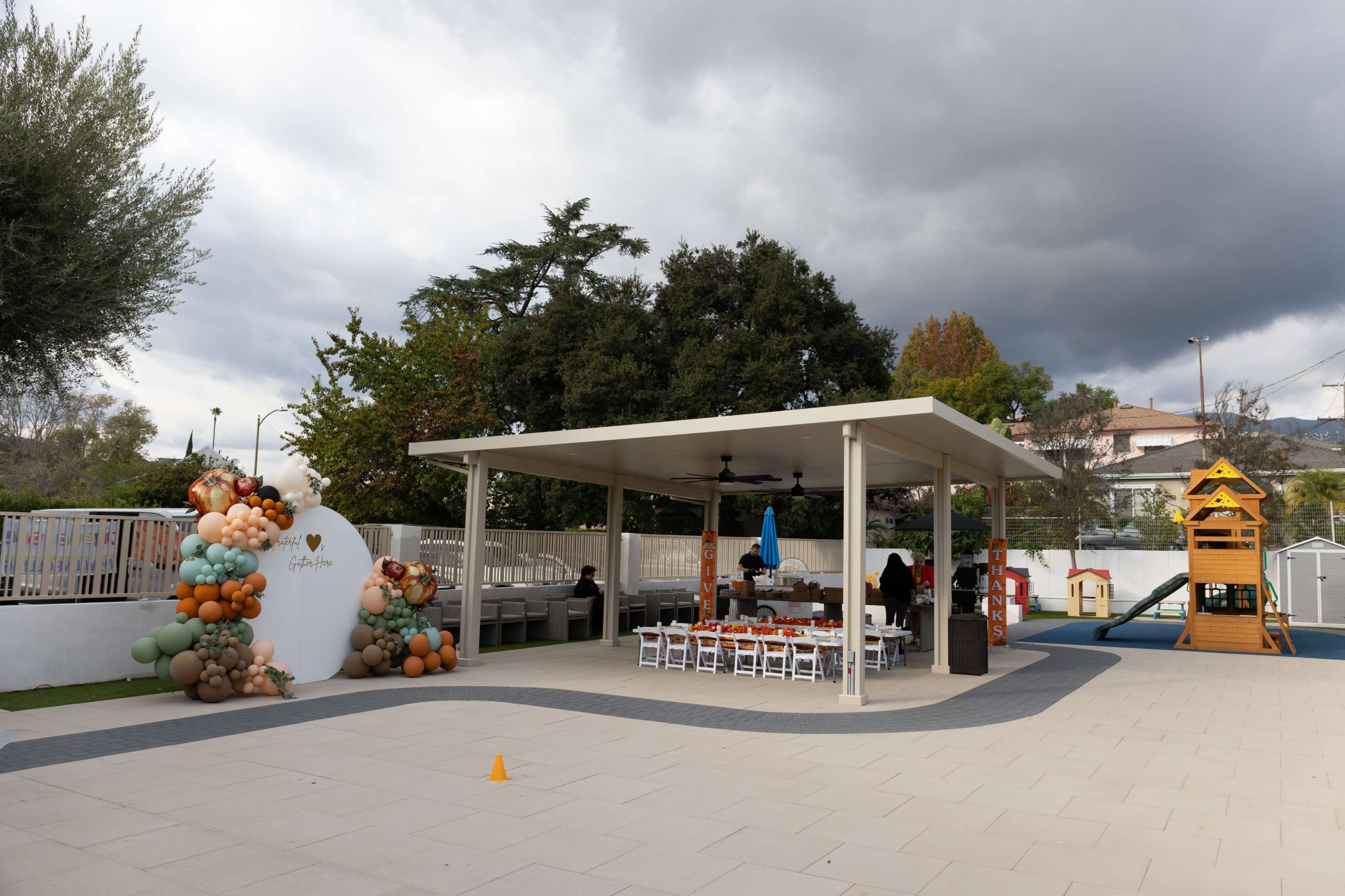 The image shows a covered outdoor area with tables and chairs, decorated with balloons, alongside a playground structure and a gray sky in the background.