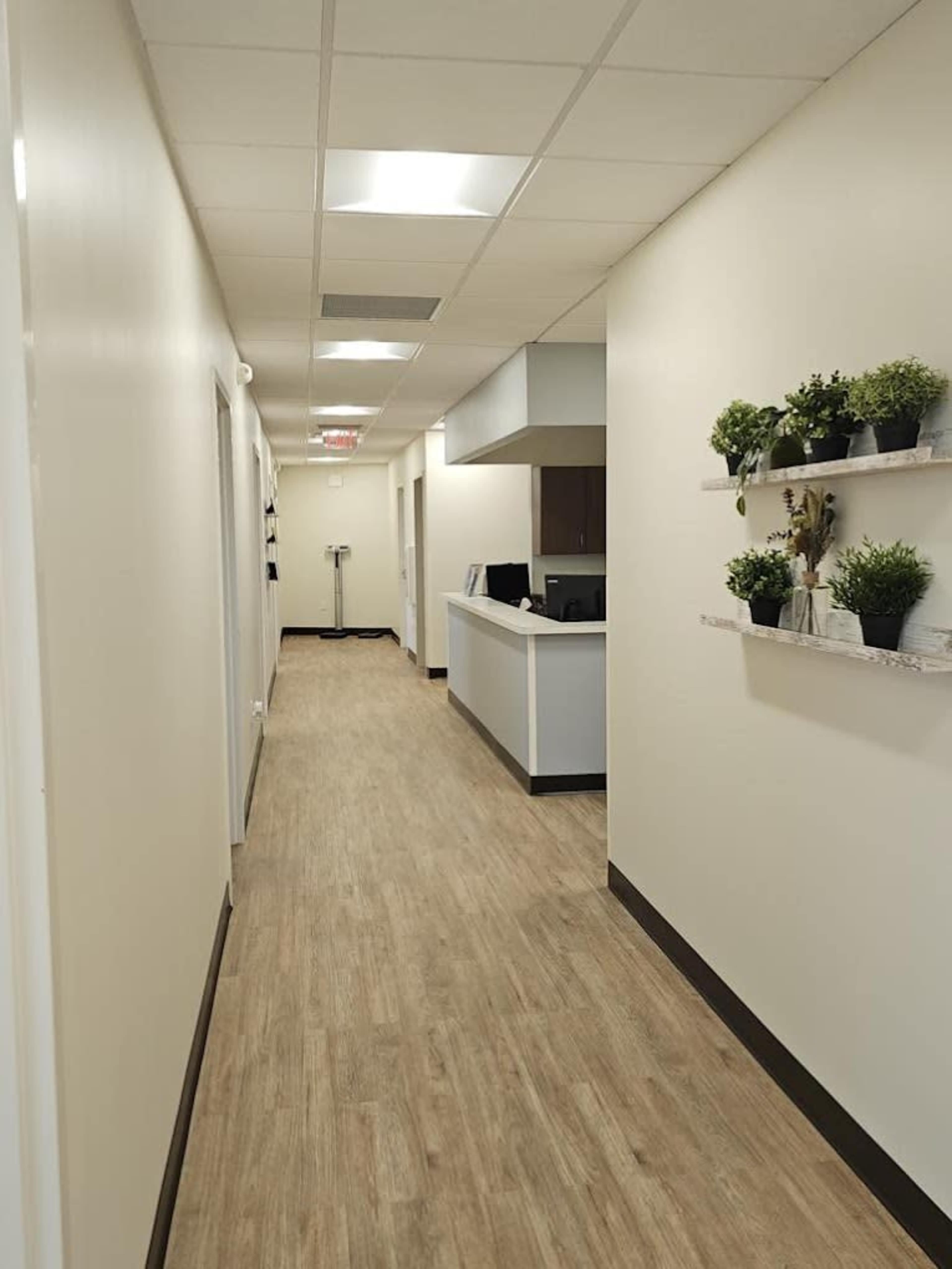 The image shows a long, well-lit hallway with light-colored walls, a reception desk at the end, and greenery displayed on shelves along one side.