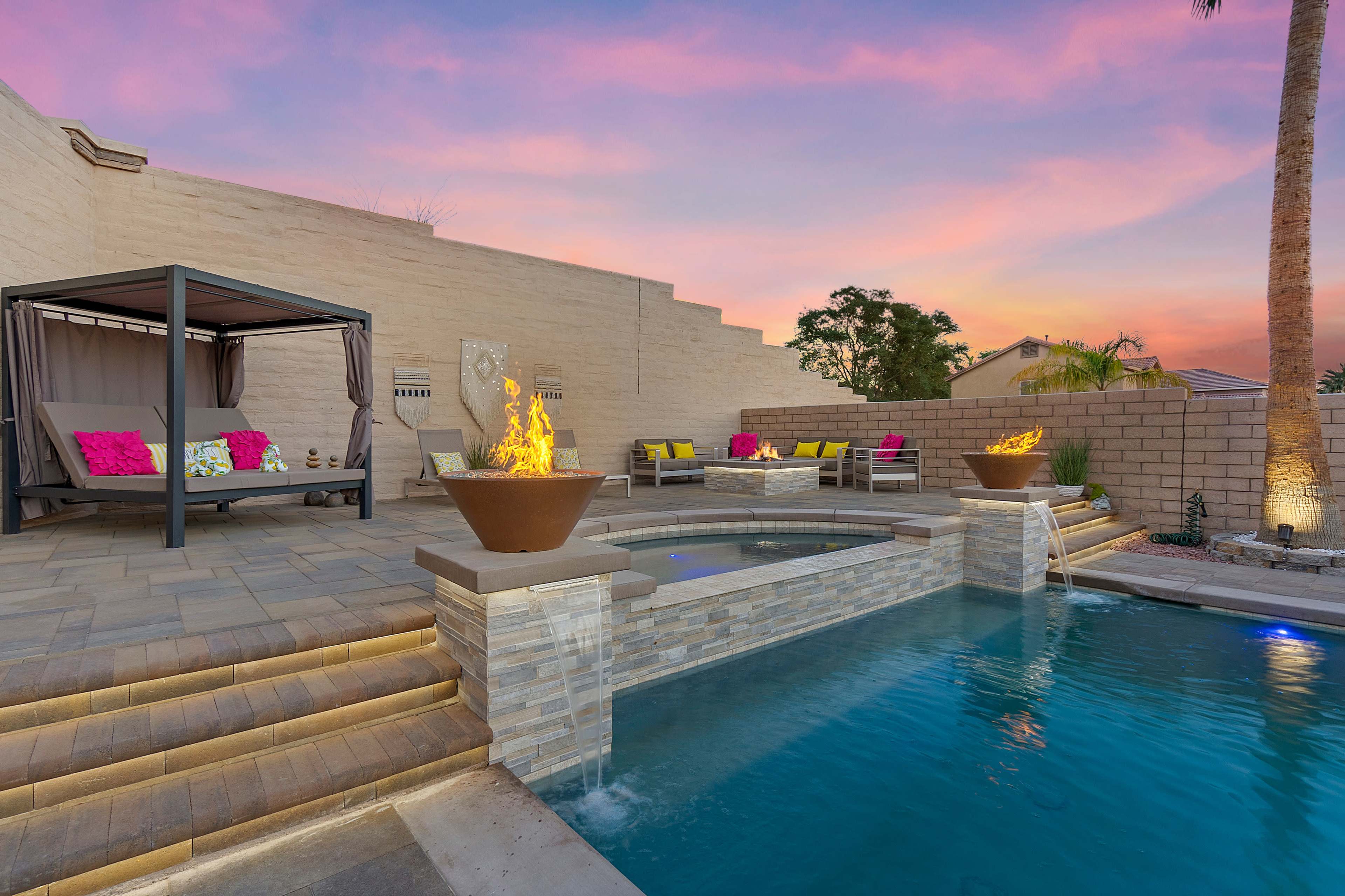 The image shows a patio area with a pool, spa, and a cabana, surrounded by landscaped walls and illuminated fire features at twilight.