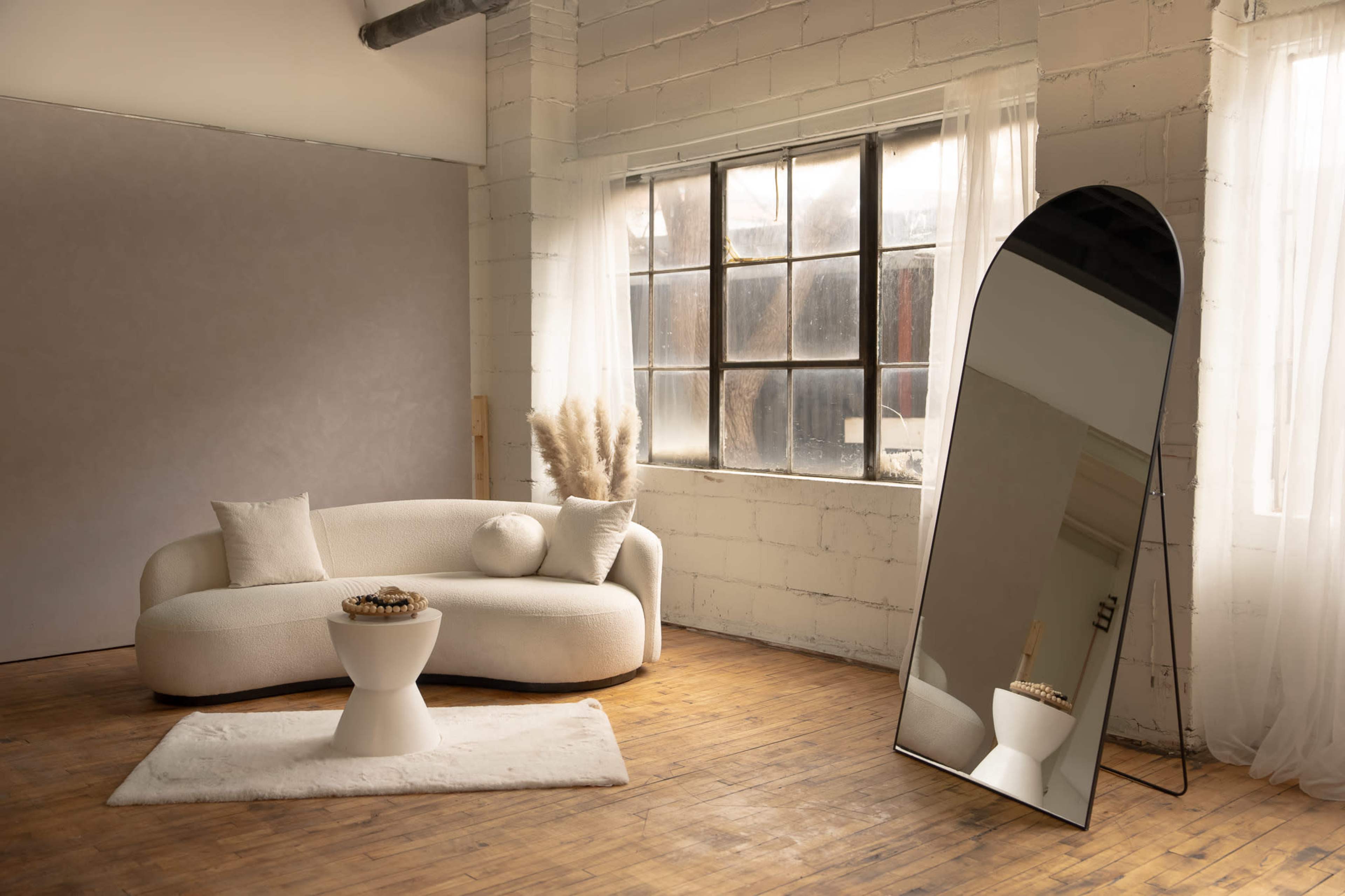 A modern, minimalistic living space features a curved white sofa, a round coffee table, and a tall, arched mirror next to a window with sheer curtains.