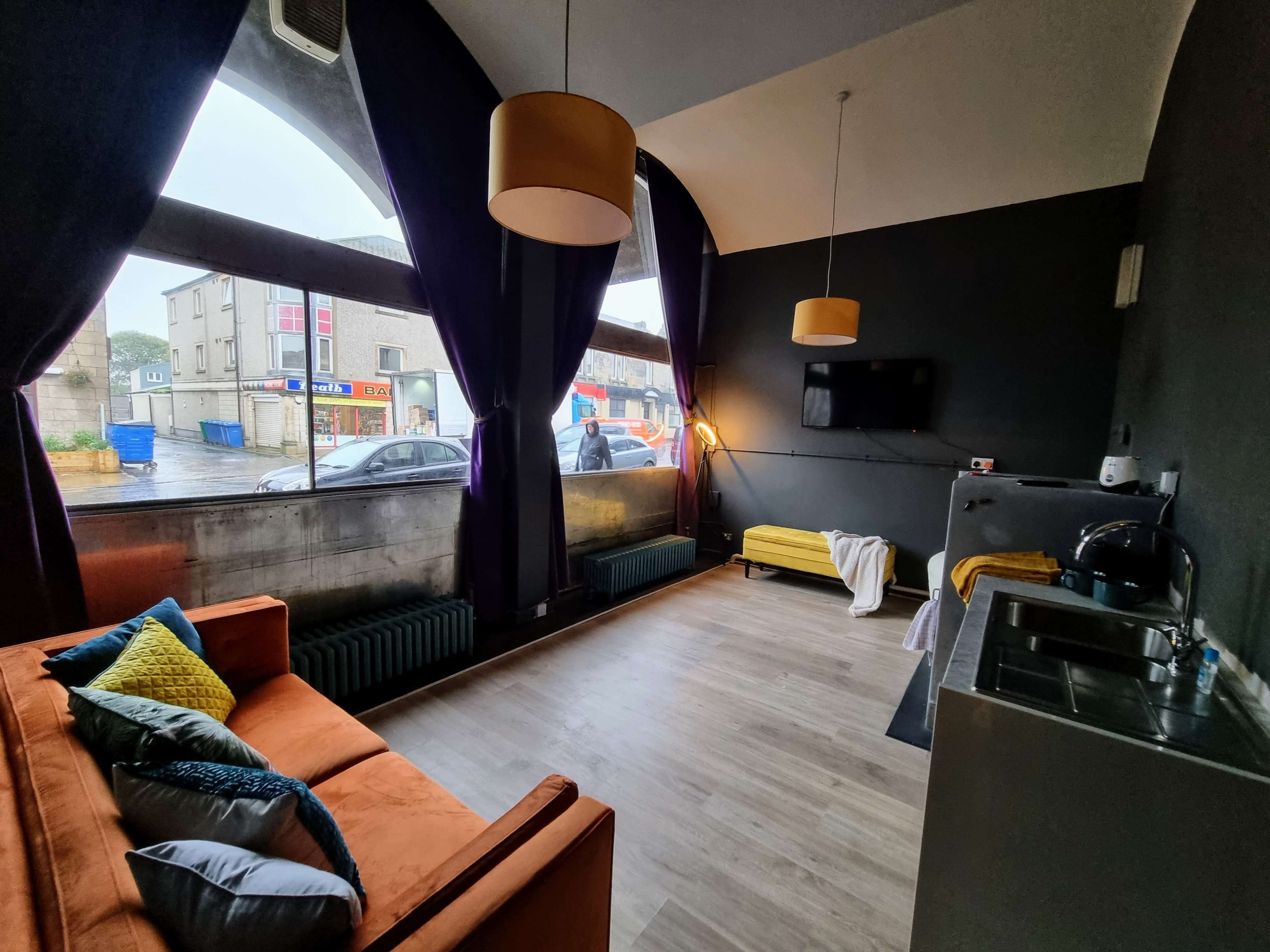 A modern interior of a room with large windows, a sofa, a kitchenette, and a view of a street outside.