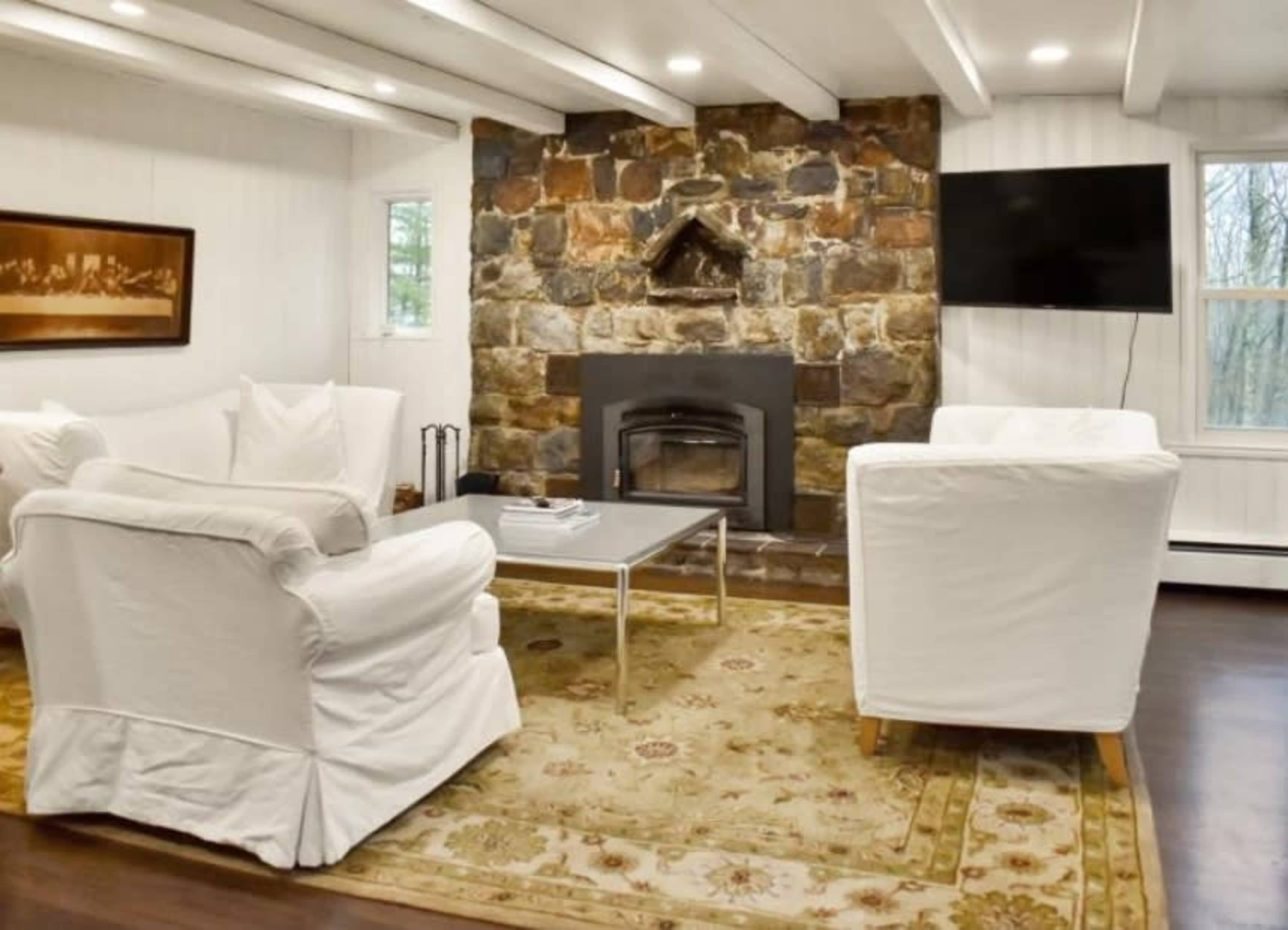 A living room features two white armchairs facing a stone fireplace, with a coffee table in front and a wall-mounted television.