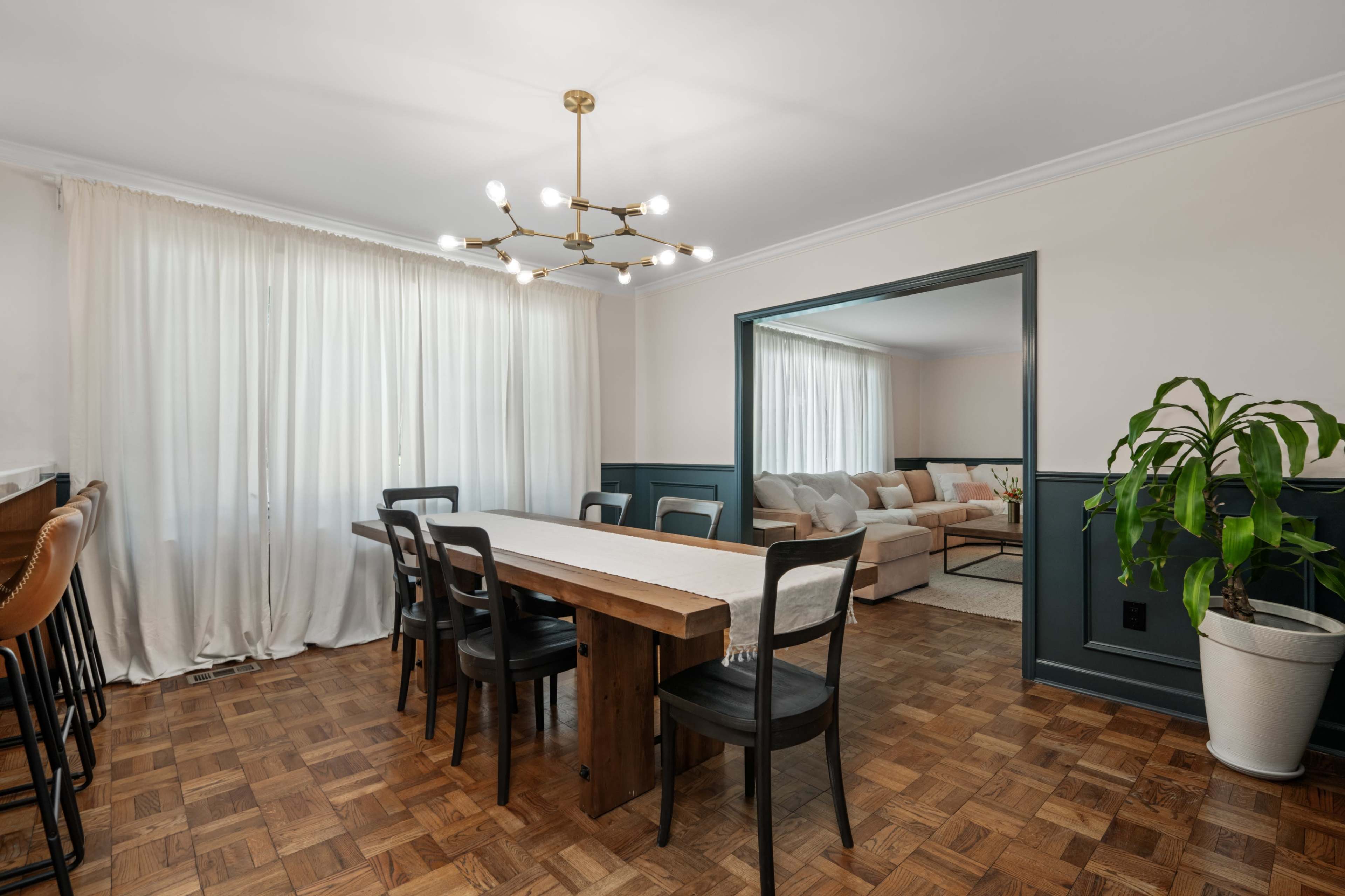 A dining room features a long wooden table surrounded by black chairs, with a chandelier above and large windows dressed in sheer curtains.